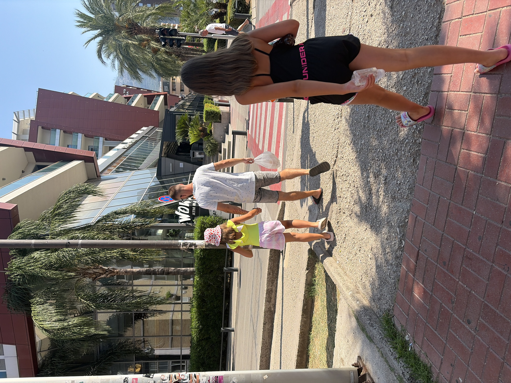 The image captures a sunny street scene in Budva, Montenegro. In the foreground, a young girl with long brown hair, wearing a black tank top with "UNDER" written in pink and black shorts, walks away from the camera. She is holding a clear plastic water bottle. To her left, a man and a younger girl walk across a crosswalk. The man, wearing a white t-shirt and grey shorts, holds a white plastic bag. The girl, wearing a yellow top and pink and white tie-dye shorts and a pink and white patterned hat, holds the man's hand. Behind them, palm trees line the street, and a modern building with glass windows and red accents stands prominently. The street is paved with asphalt and concrete, with a red brick walkway in the foreground. A traffic light and a pole with a warning sticker are visible on the left. The sky is clear and blue.