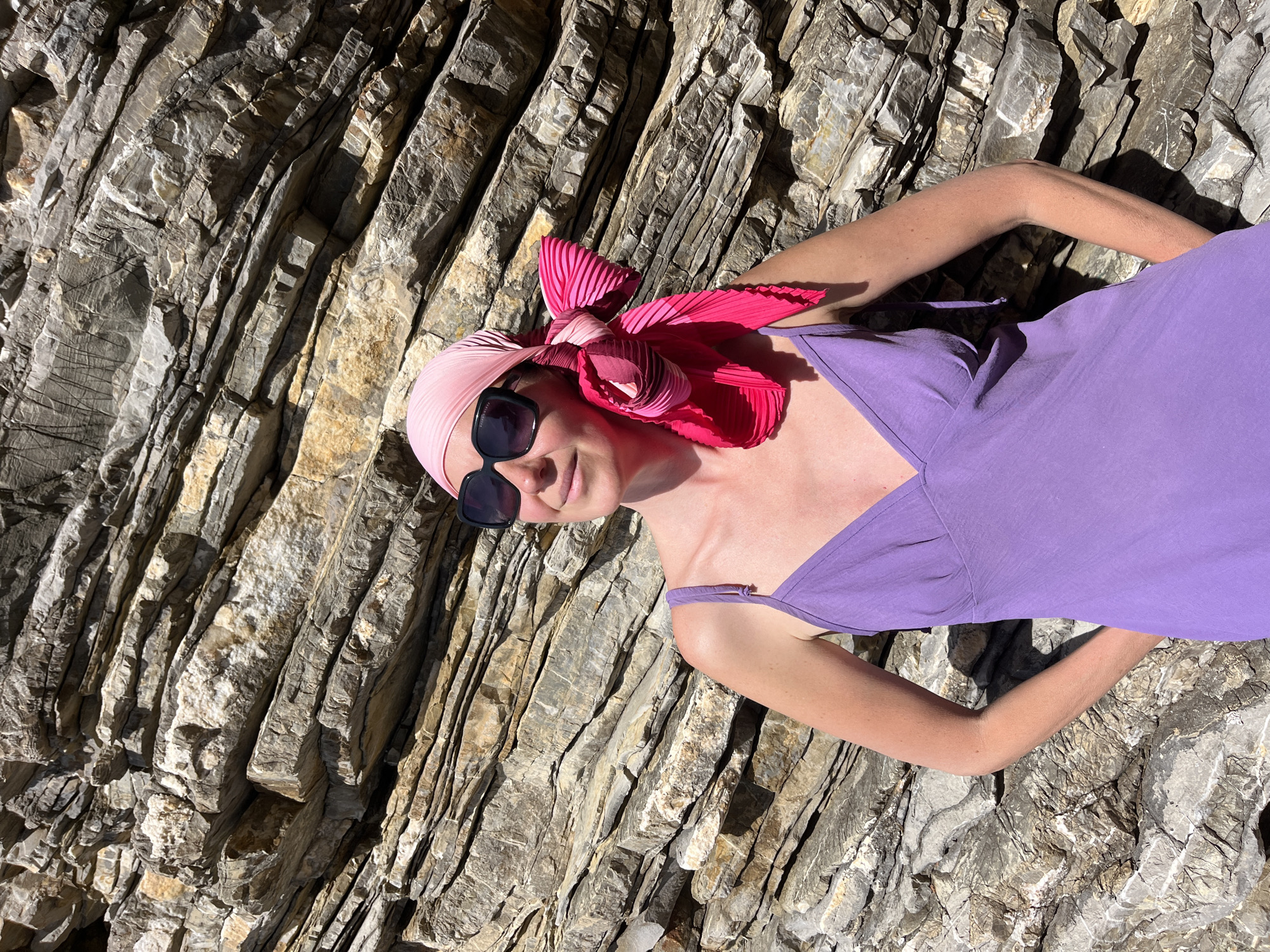 A woman lies on a textured rock formation. She wears a lavender dress, sunglasses, and a pink headscarf with a pleated pink bow. The rock is a stratified grey-brown color, with crevices and shadows creating visual texture. The scene is brightly lit, and the location is near Budva, Montenegro, as evidenced by the distinctive rock formations common to that area.