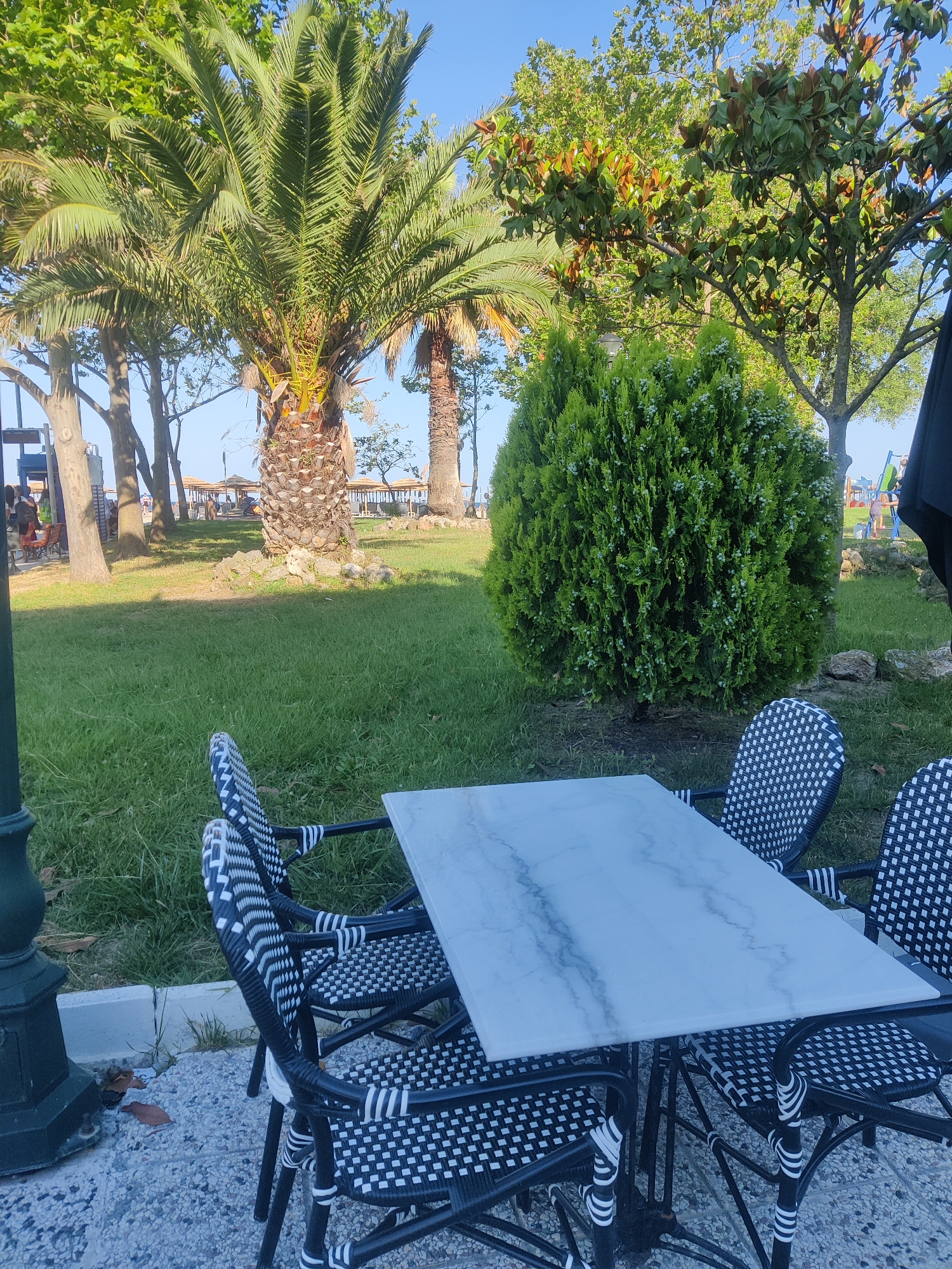 An outdoor table with four black and white patterned chairs is set on a pebbled patio in front of a lush green lawn. A white marble-patterned table sits at the center of the arrangement, with chairs positioned around it. In the background, palm trees, manicured bushes, and various other trees create a vibrant natural setting under a clear blue sky. Further in the distance, a beach area with umbrellas and people can be seen. The scene appears to be a cafe or restaurant patio located in Katerini Municipality, Greece, likely near a beach resort or promenade.