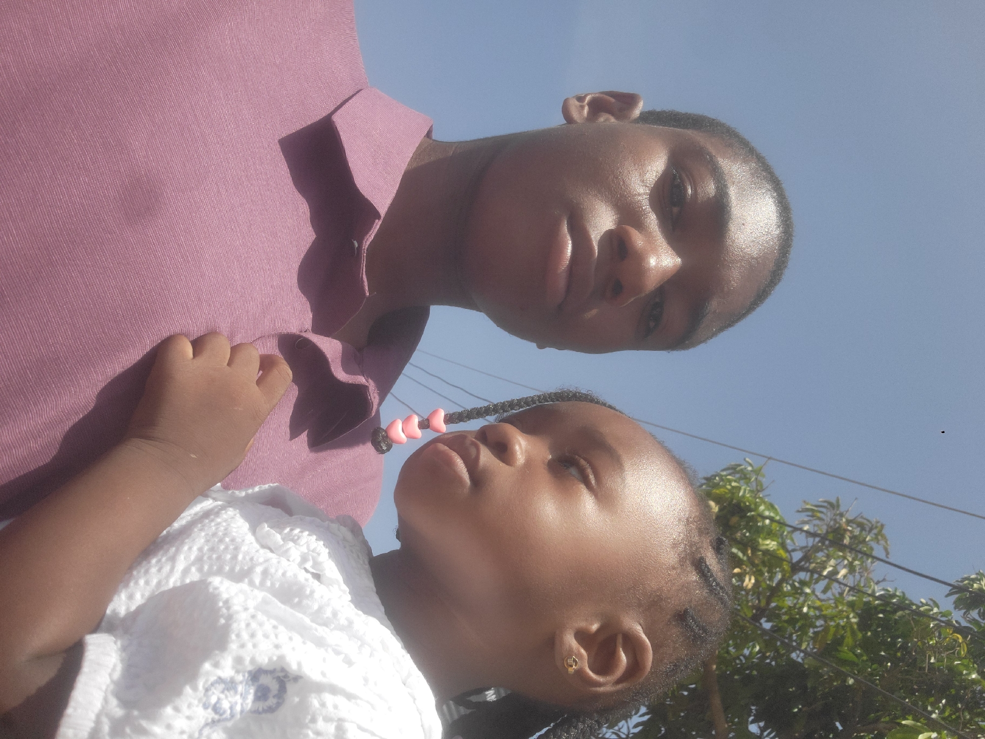 A low-angle medium shot depicts two individuals. The individual closest to the camera appears to be a young child with braided hair adorned with beads. They are wearing a white textured garment. The second individual, seemingly an older male, stands behind the child, wearing a collared purple shirt. They are positioned against a clear blue sky with partial foliage in the background. The photograph seems to be taken outdoors in daylight. There is no explicit evidence within the image to corroborate its location as Jos, Nigeria.