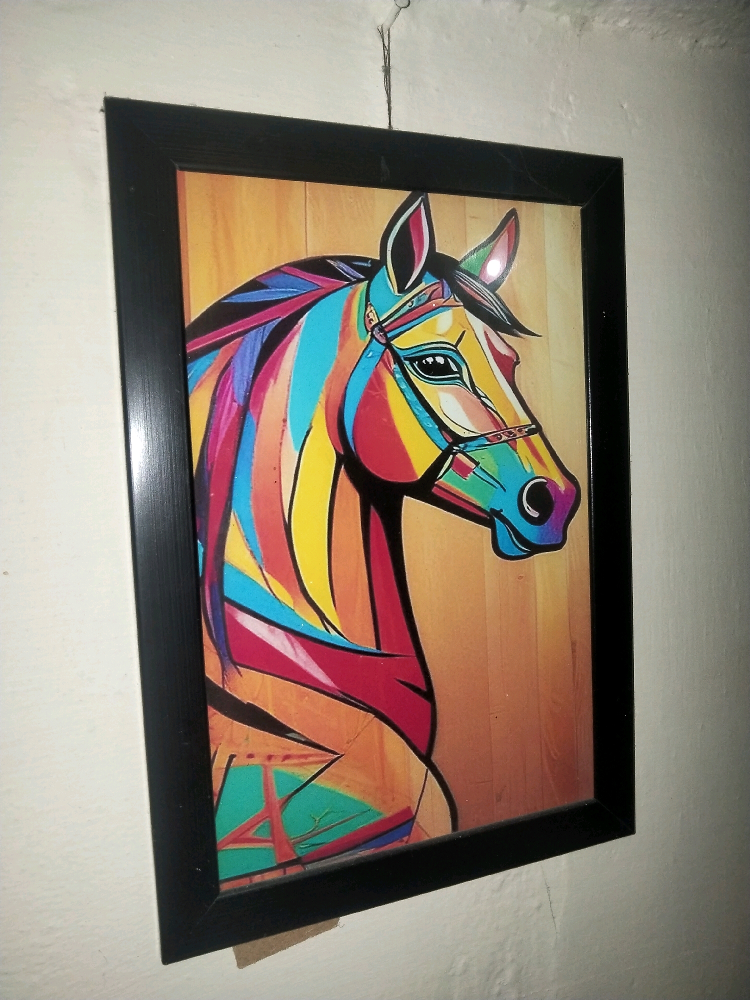 A framed painting of a colorful, abstract horse is hanging on a white wall. The painting depicts the horse's head and neck in profile, rendered with bold lines and vibrant hues of red, blue, yellow, and green. The background of the painting is a warm, light brown, possibly representing wood grain. The frame is black and appears to be made of wood or plastic. The painting is suspended by a thin wire attached to a small hook at the top. The wall itself is textured and appears to be slightly uneven. There is no visible indication of the specific location within Haripur, Pakistan.