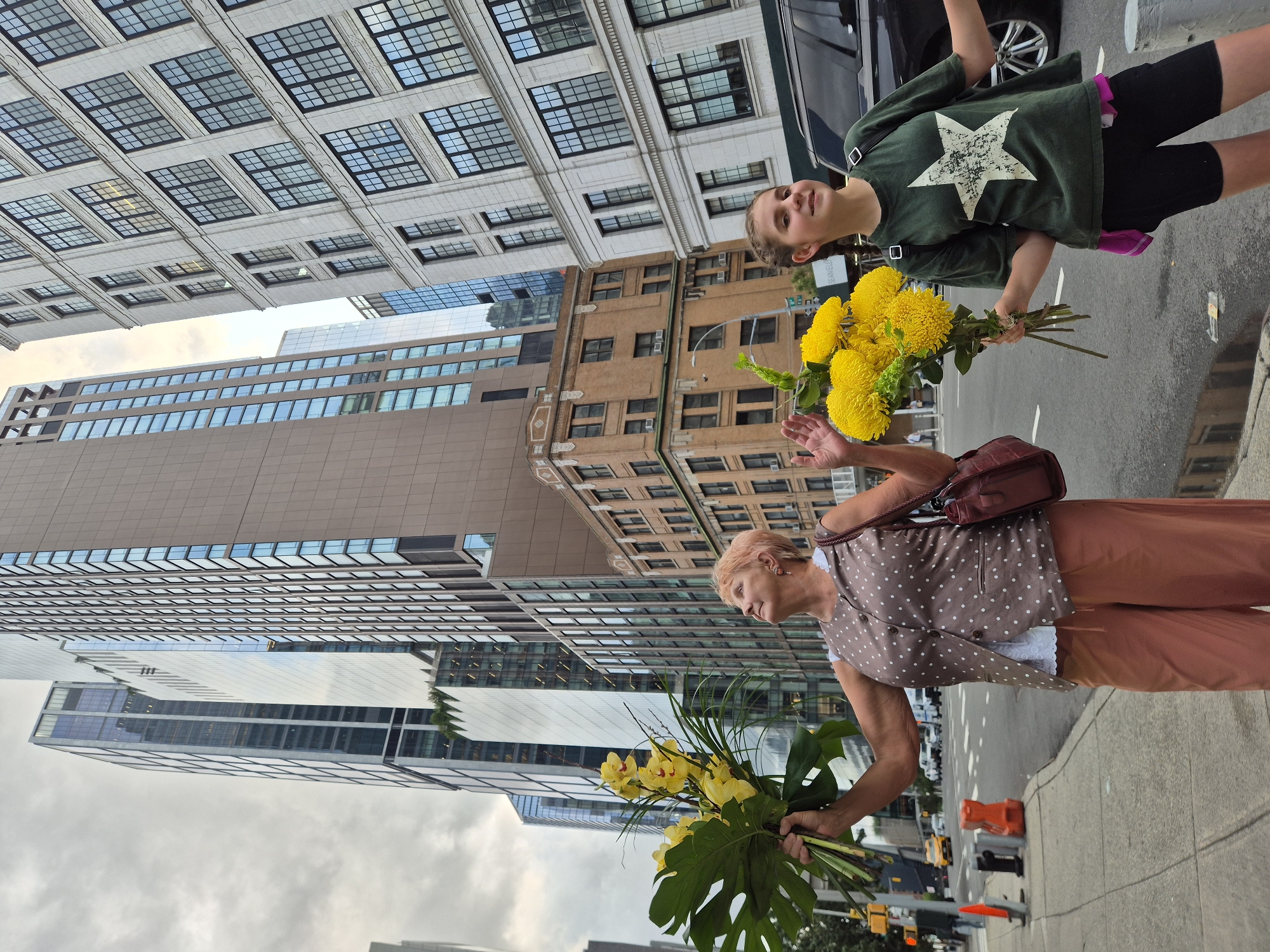 Two people, an older woman and a younger girl, are holding bouquets of yellow flowers while standing on a sidewalk in New York City. The woman, on the left, has pink hair and is wearing a brown polka-dot vest over a white shirt and peach-colored pants. She is looking up and to her left, with a large bouquet of yellow orchids and monstera leaves in her left hand. The girl, on the right, has braided brown hair and is wearing a green t-shirt with a white star graphic, black shorts, and a pink garment tied around her waist. She is smiling and holding a bouquet of yellow chrysanthemums. They are standing in front of tall buildings with many windows, characteristic of a dense urban environment. A street with traffic, including cars and traffic signals, is visible behind them. The sky is overcast. The architecture of the buildings suggests a location in Manhattan.