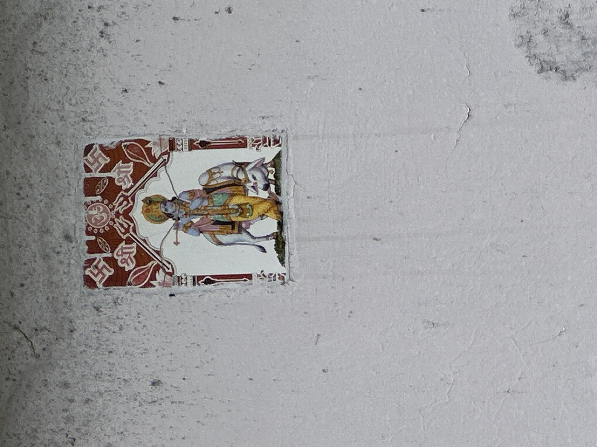 The image shows a religious decal adhered to a textured, light-colored wall. The decal depicts a Hindu deity, likely Krishna, in a stylized, colorful illustration. The deity is shown with a blue complexion, wearing ornate clothing and a headdress, and is holding a bow and arrow. To the right of the deity, a white cow with a decorated hump and head is depicted, symbolizing Nandi, the bull of Shiva, or a sacred cow in Hinduism. The decal is framed by decorative elements that include swastikas and floral motifs. The wall itself appears weathered and shows signs of age and possibly minor damage, such as small cracks and an uneven surface texture. The scene is devoid of any human interaction or activity, with the decal being the sole focal point on the otherwise plain wall. The location is identified as Haini, India.