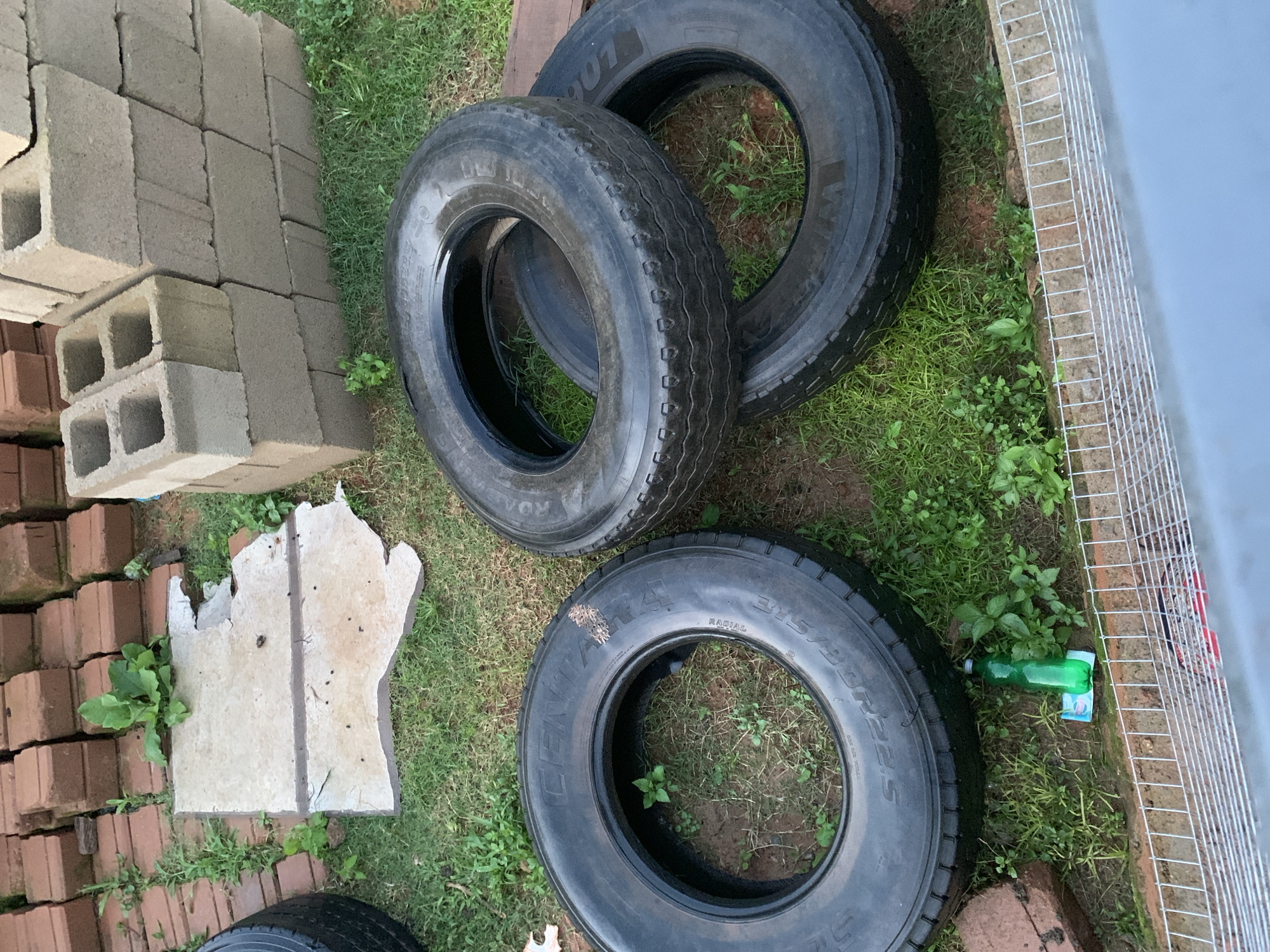 In an outdoor setting, likely a backyard in an unknown location, several objects are present. Three large tires are the most prominent subjects, arranged on a patch of grass. One tire is on the left side of the frame, lying flat and open. Another tire is placed on top of another tire, also lying flat, in the center of the image. The third tire is partially visible beneath the tire in the center. Behind the tires, a stack of reddish-brown bricks is visible, as well as a stack of concrete blocks. To the left of the tires, a broken piece of concrete pavement lies on the grass, with a small plant growing out of it. In the bottom right corner of the image, a wire fence runs across the frame. A green plastic bottle and a white object are on the grass near the fence. The ground is covered in grass and dirt. The lighting suggests it is daytime.