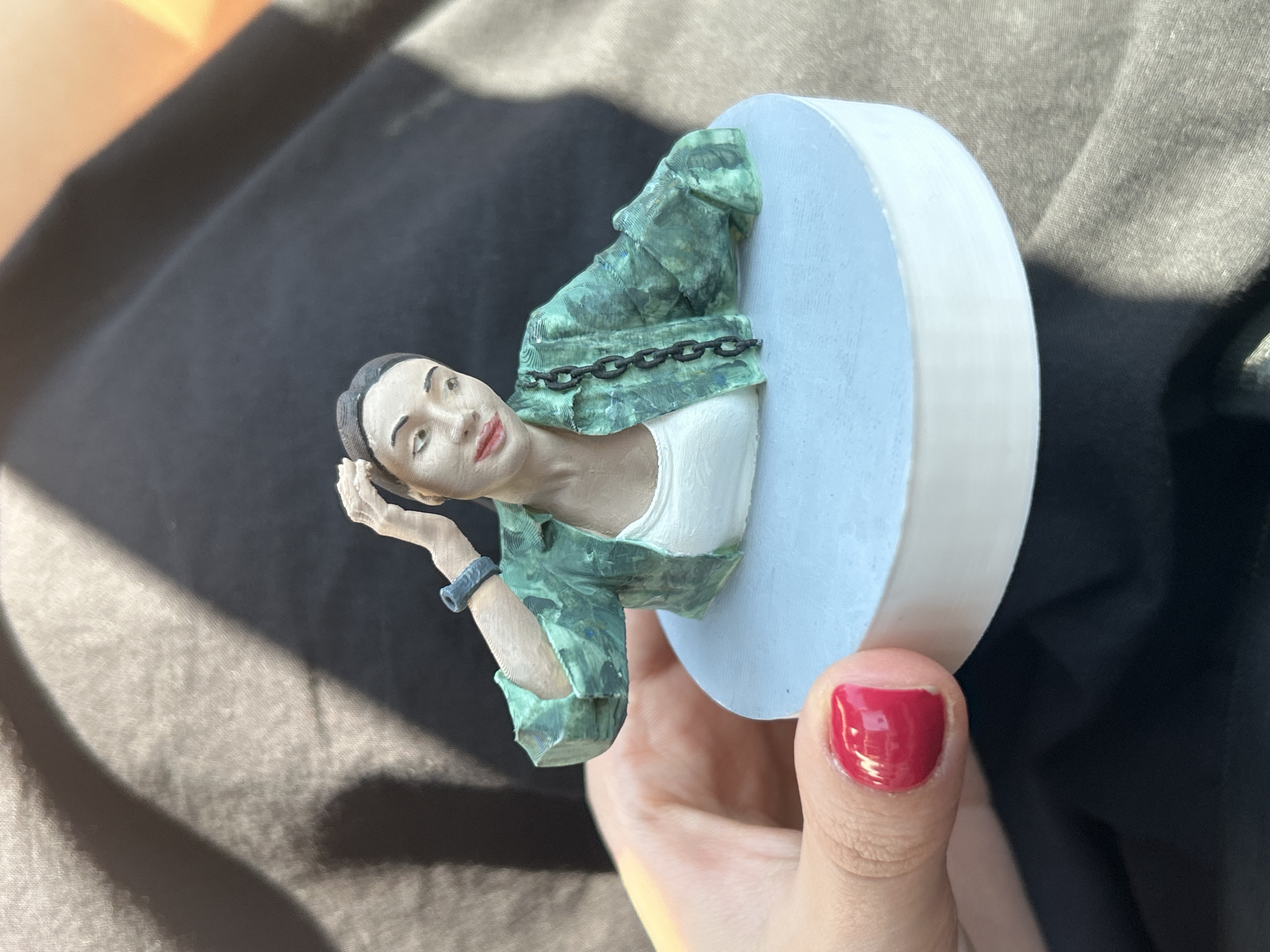A 3D printed figurine of a woman is held in a person's hand, with a dark fabric background. The figurine depicts a woman with dark hair pulled back, wearing a green patterned jacket over a white top. She has a chain across her jacket and a blue bracelet on her left wrist. Her right hand is raised to her head. The figurine stands on a circular base that is light blue on top and white on the bottom. The hand holding the figurine has a fingernail painted bright red. The background is out of focus and appears to be dark clothing with some light shadows casting across it. There are no identifiable landmarks or contextual clues to suggest a location in Sofia, Bulgaria.