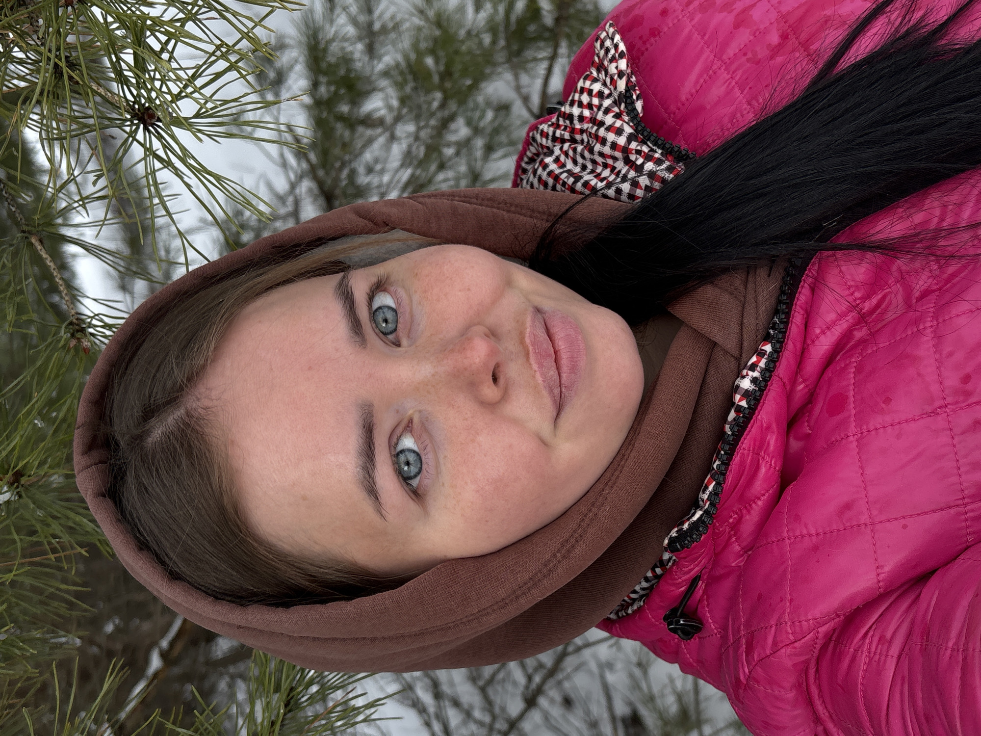 A young woman with fair skin and light blue eyes, wearing a brown hoodie and a pink quilted jacket with a black and white checkered trim, is looking at the camera. She has dark hair. The background includes pine tree branches and a snowy landscape, suggesting an outdoor winter setting. The photograph was taken in a way that makes the composition seem tilted. The location is Русский Вожой, Russia.