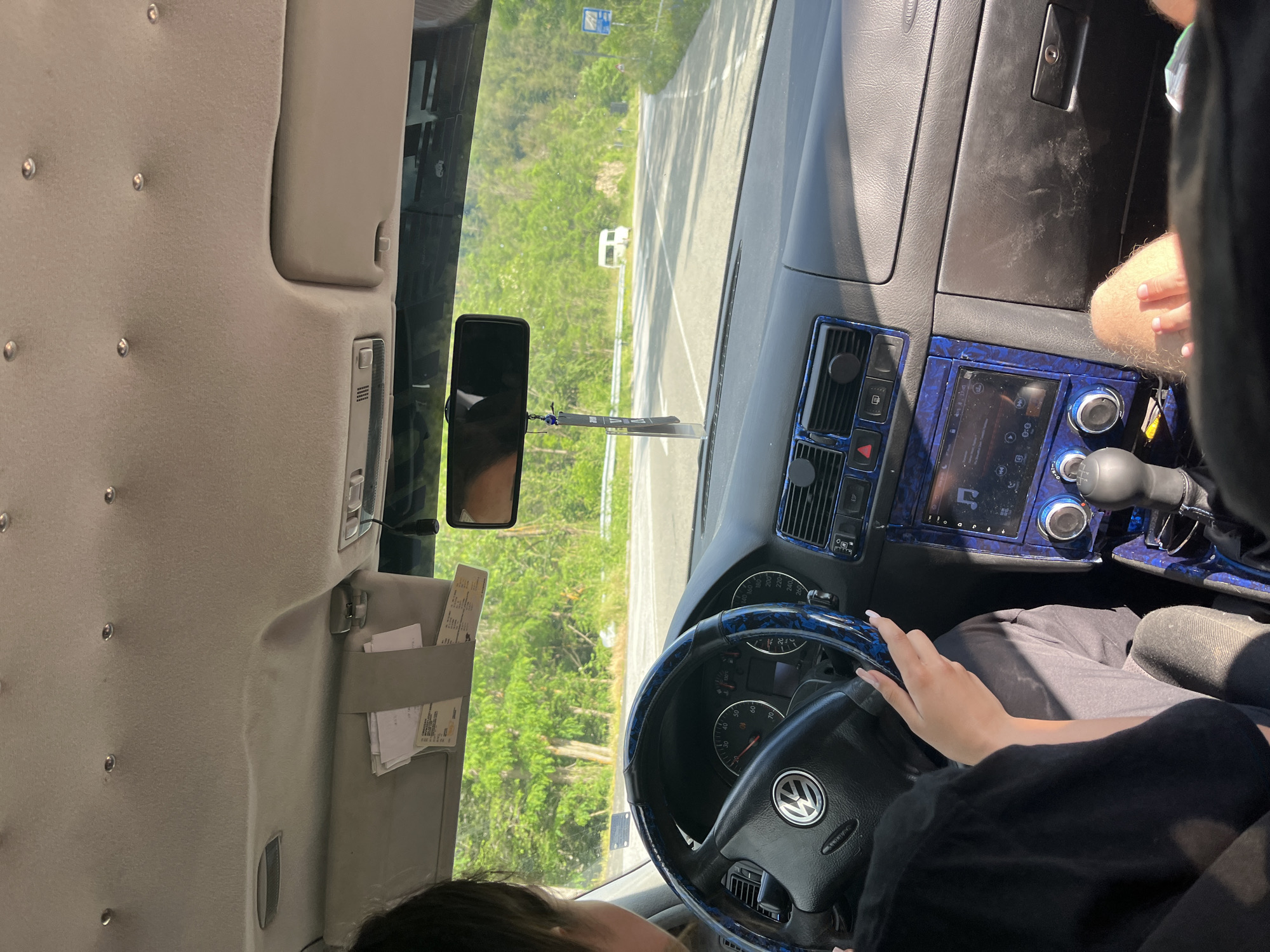The image shows the interior of a car, looking forward. A woman is driving, with her hands on the steering wheel. The dashboard has a blue, patterned trim. Outside the car, there is a road with trees and hills in the background. A white van is visible in the distance. The scene appears to be captured on a sunny day, likely in the city of Leshtak, Bulgaria.
