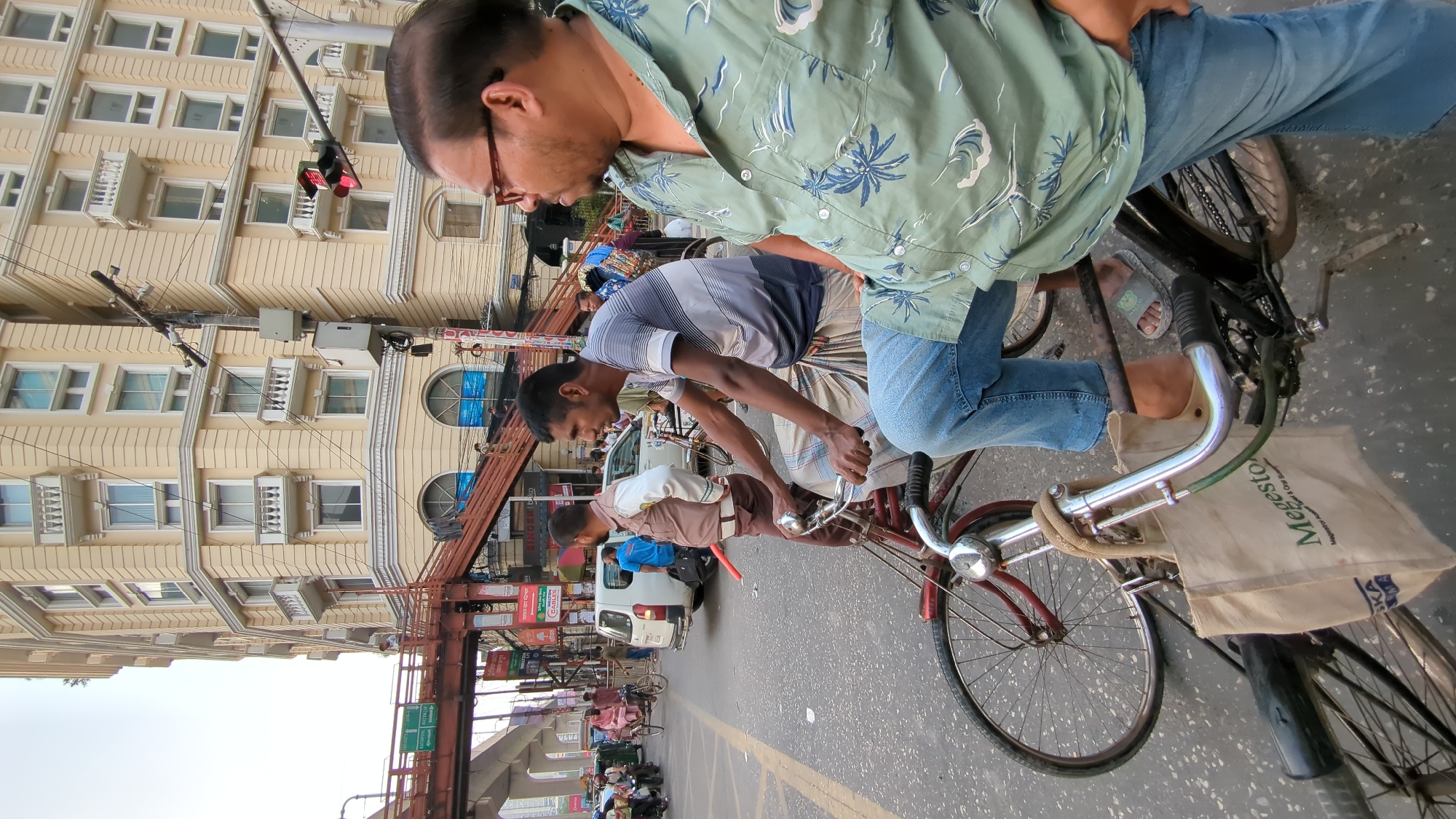 The image captures a bustling street scene in Dhaka, Bangladesh. Three men are riding bicycles in the same direction on a busy city street.
The first man, closest to the viewer, is wearing a light green short-sleeved shirt with a blue floral pattern and blue jeans. He is riding a red bicycle with a tan bag hanging off the front. The middle man is wearing a white striped shirt and lungi. The third man is wearing a light brown shirt.
Overhead electric lines and traffic signs crowd the upper portion of the frame. A beige building with symmetrical windows stands on the left. Cars and pedestrians are visible in the background, indicating a crowded urban environment.