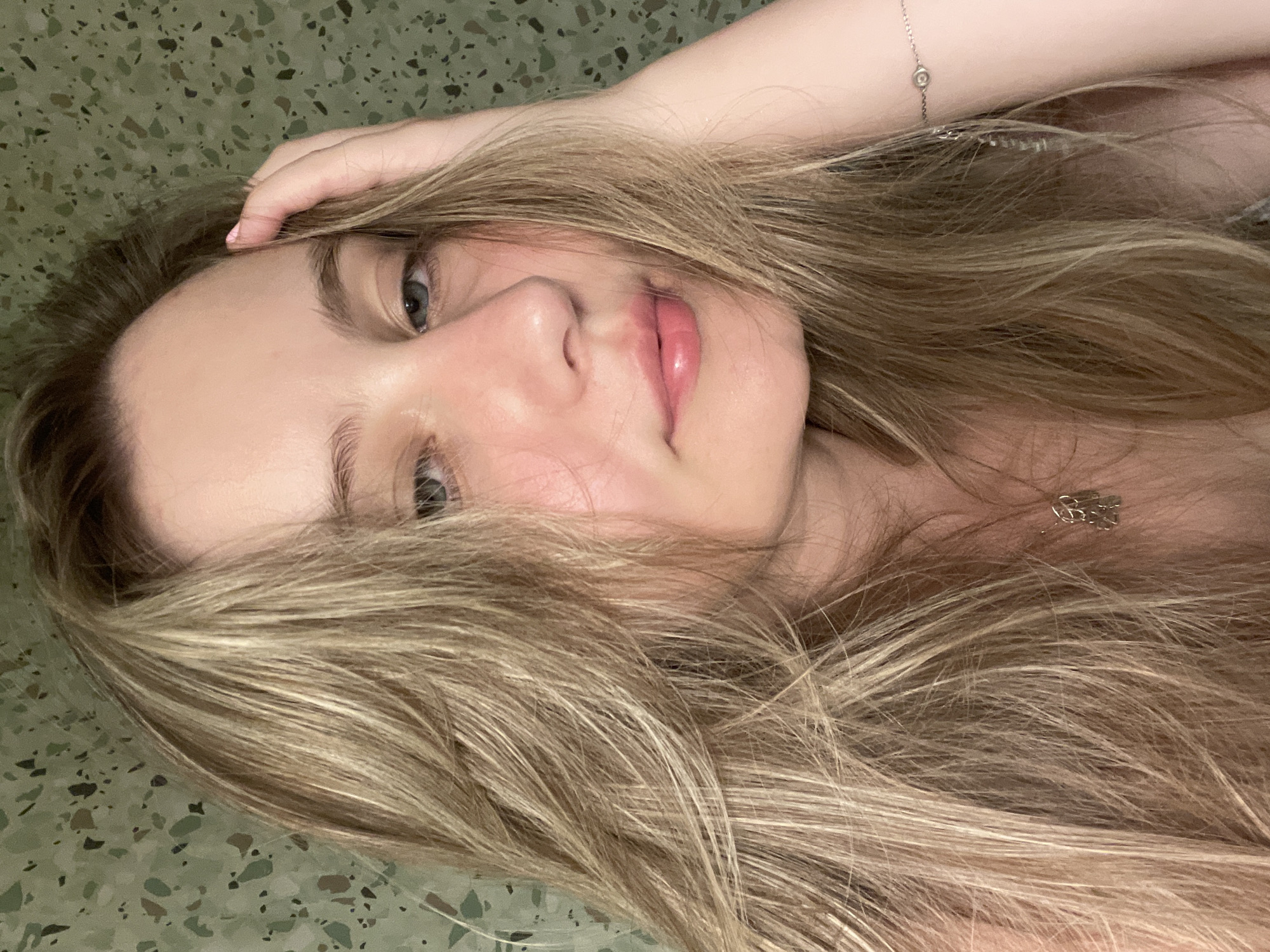 A young woman with blonde hair is resting her head on her hand, looking at the camera. She is wearing a necklace and a bracelet. The background is a green wall with a speckled pattern.