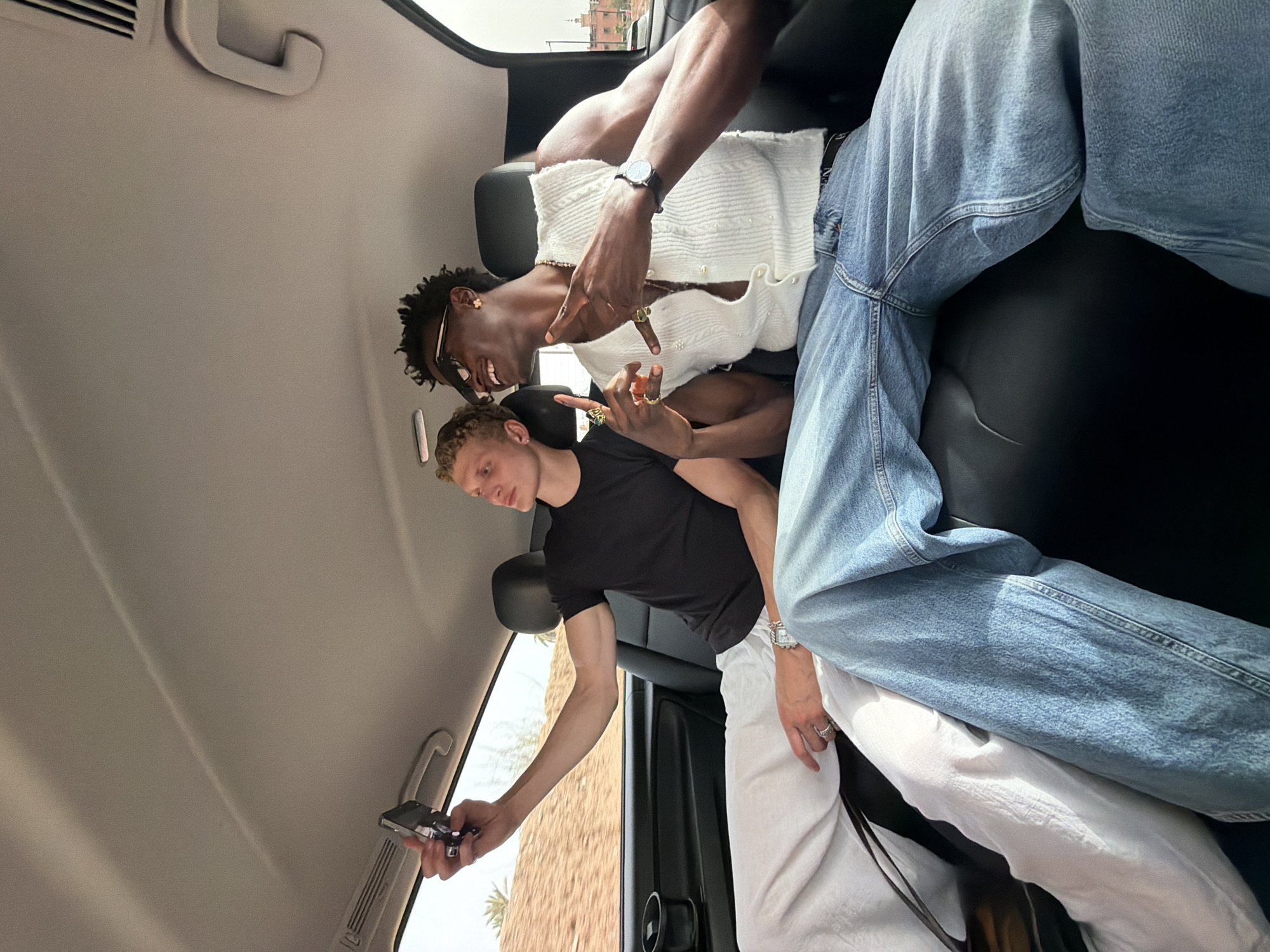 Two young men are seated in the back of a vehicle, possibly a van or SUV, in Marrakesh, Morocco. The man on the left, with blonde curly hair, is wearing a black t-shirt and white linen pants. He is holding a smartphone in his right hand, taking a selfie. He wears a silver watch and rings on his fingers. The man on the right, with dark curly hair, is wearing sunglasses, a white sleeveless knit vest over a dark top, and blue jeans. He is smiling and making a hand gesture with his right hand. He has rings on his fingers and a necklace. The interior of the vehicle is visible, including black leather seats and the ceiling. Through the window on the left, a landscape with sand and palm trees can be seen, suggesting an arid environment. In the background, a distinctive red building with architectural details, characteristic of Marrakesh, is visible. The lighting suggests it is daytime, possibly late afternoon.