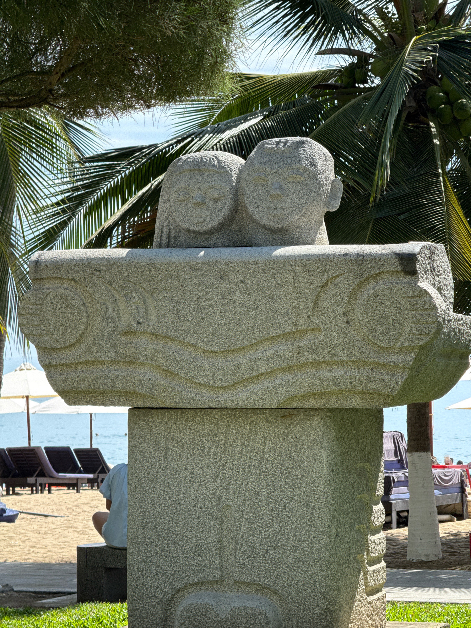 In Nha Trang, Vietnam, a carved stone sculpture of two faces, possibly children, is positioned centrally in the frame, overlooking a beach scene. The sculpture is made of speckled granite and rests on a pedestal, with a stone slab featuring carved wave-like patterns extending from its base. In the background, palm trees with coconuts provide a tropical canopy, their fronds reaching towards the bright blue sky. The beach is visible with lounge chairs and umbrellas, and the ocean stretches out to the horizon. A person, dressed in a light blue shirt and white shorts, sits on a stone bench to the left of the sculpture, facing away from the camera and towards the beach. The overall setting appears to be a resort or public space near the coast.