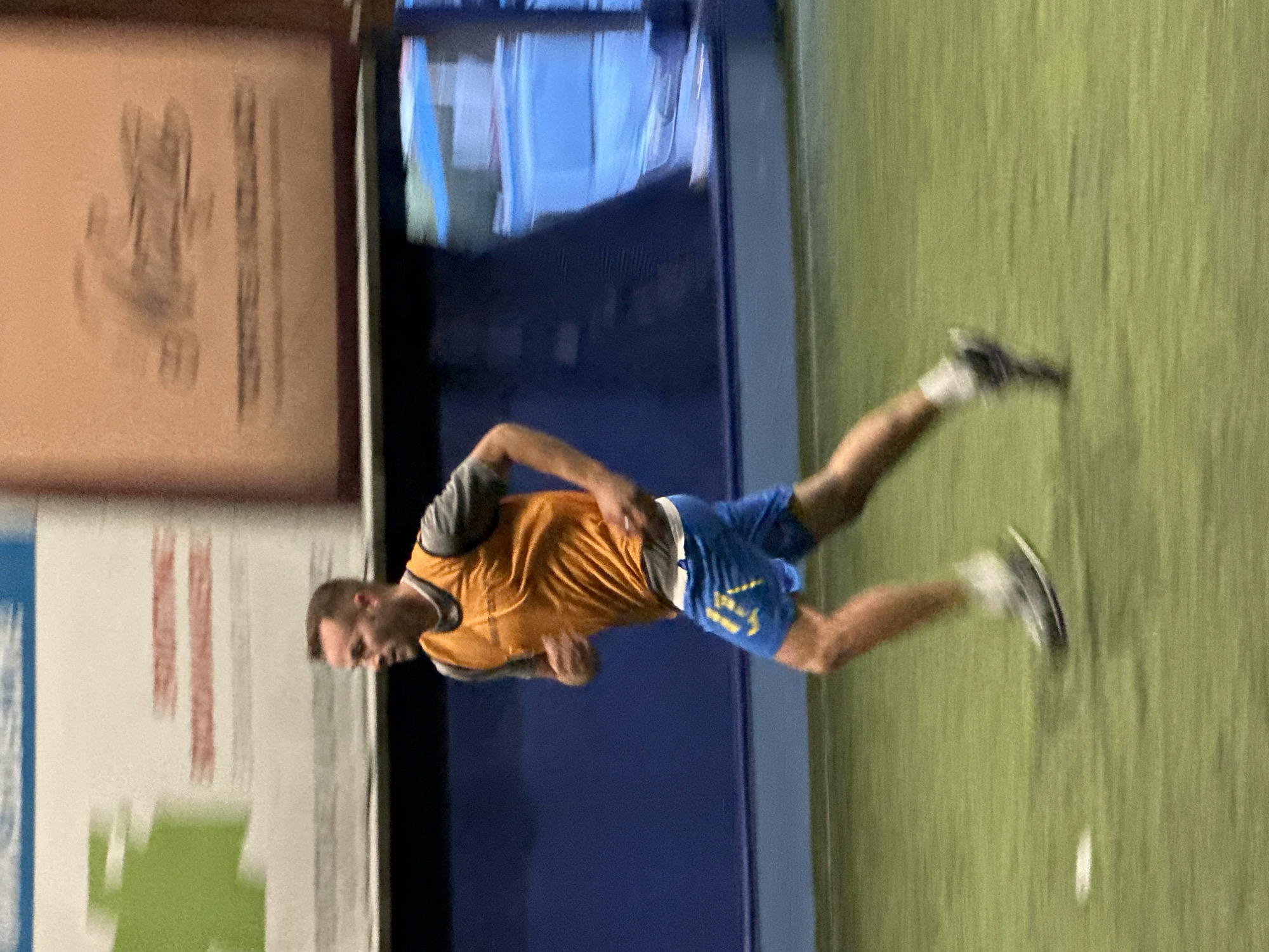 A man in an orange jersey and blue shorts is running across a green surface. The background includes a blue wall, a framed picture on a wall, and possibly a window. The location within the city of Budva, Montenegro, cannot be determined from the image.
