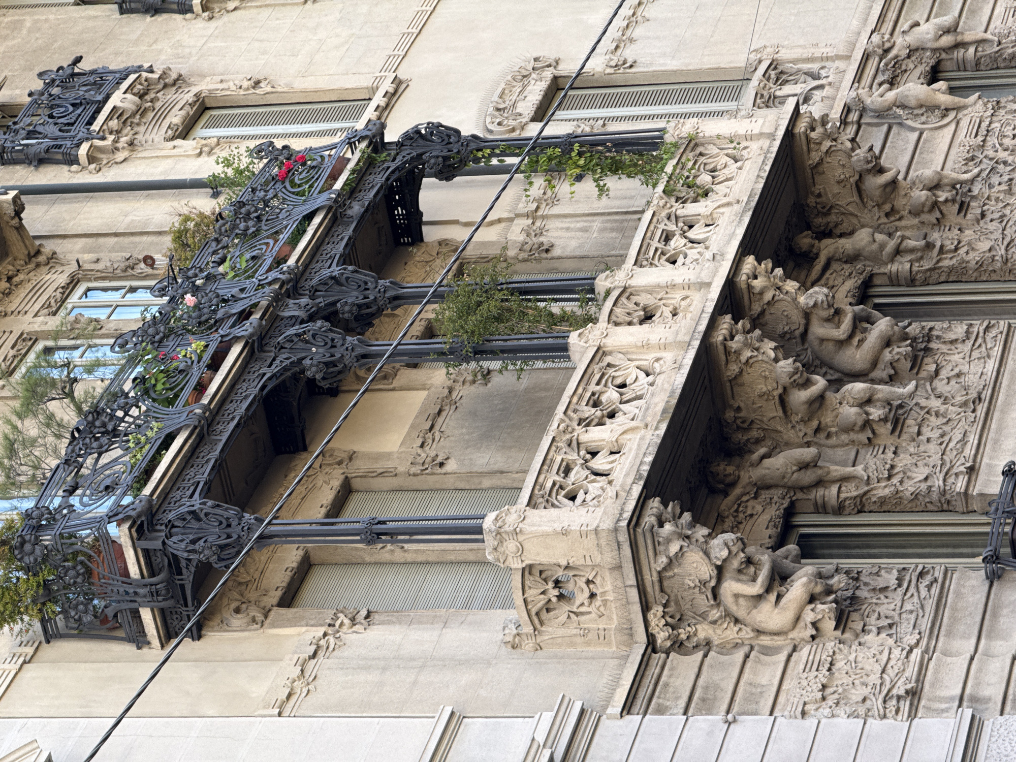 The image shows a section of an ornate building facade in Milan, Italy. The building has a pale, beige-colored exterior. The architectural style is decorative, featuring extensive sculptural details. On the left side of the image, there is a wrought-iron balcony filled with plants and flowers. A wire diagonally crosses the front of the building. To the right, the building has a column of deeply carved relief sculptures, depicting reclining figures, possibly cherubs or other allegorical beings. Windows with horizontal blinds are visible between the sculptural elements. The architecture suggests a historic building, potentially dating from the late 19th or early 20th century. The photograph is taken from a low angle, looking upwards. The sky is reflected in one of the windows.