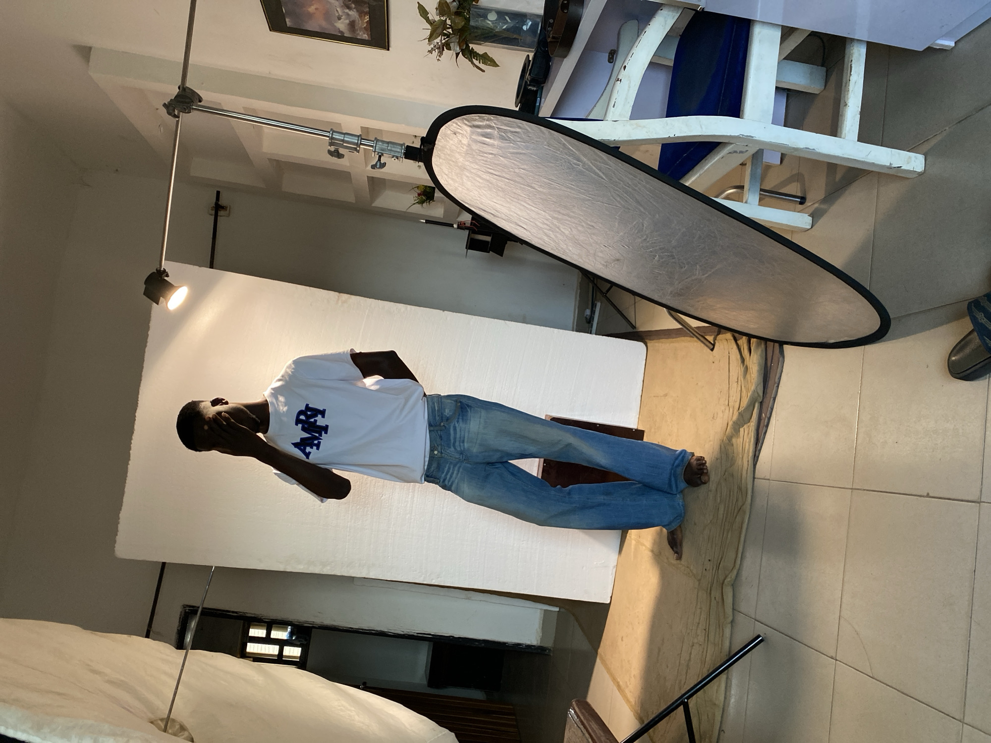 The image captures an indoor photography setup, likely a studio, in Bukuru, Nigeria. A dark-skinned young man, serving as the model, stands barefoot against a large white backdrop. He is dressed in a white short-sleeved t-shirt and light blue jeans. His right hand covers part of his face, suggesting either a deliberate pose or an attempt to shield his eyes from the bright artificial light. On his t-shirt, stylized blue letters resembling "AMRI" are visible.
The scene is equipped with professional photography lighting. A spotlight, mounted on a stand, illuminates the model and the backdrop from the left. To the right of the model, a large, oval-shaped silver reflector stands on its own support, positioned to bounce light. The floor is tiled with light-colored squares, and a beige cloth or rug is laid out beneath the model's feet. In the background, a distressed white wooden chair is visible, along with a framed picture and a plant in a vase, suggesting a lived-in or adaptable studio space. The overall environment indicates a photoshoot in progress.