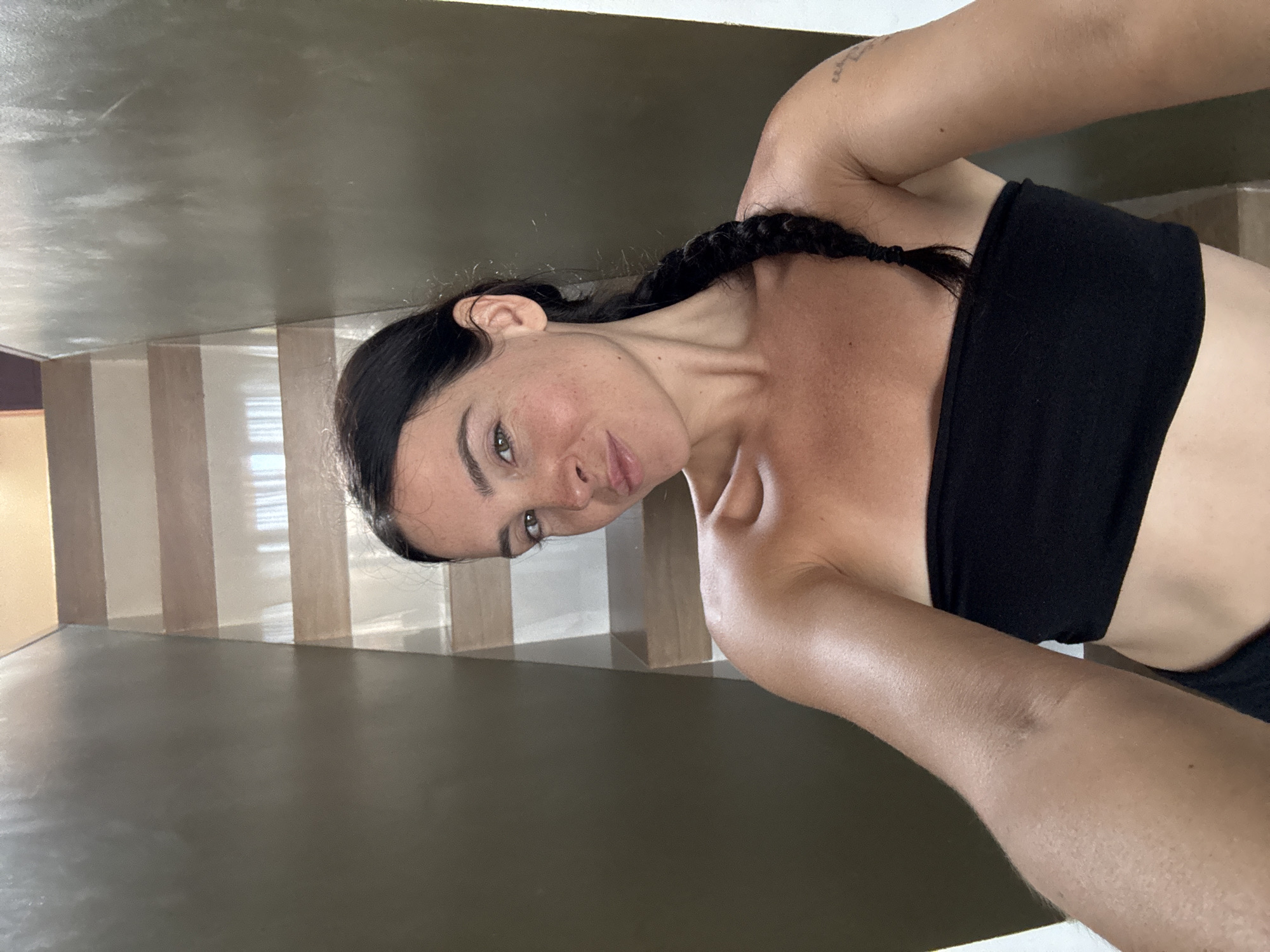 A woman with dark hair braided down her back and wearing a black bandeau top poses for a selfie. She is looking at the camera with a slight smile. Her skin is tanned, and she has a smattering of freckles across her face and shoulders. A small tattoo is visible on her upper left arm.
The background appears to be an interior space with a staircase. The walls are a mix of earthy tones and lighter stripes, suggesting a modern, possibly minimalist design. The lighting is natural and soft, hinting at daytime. The overall mood is relaxed and casual. The location is identified as North Kuta, Indonesia.