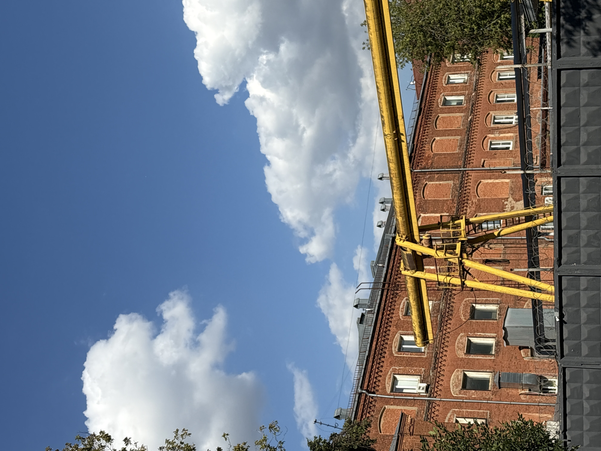 This is a photograph taken on a sunny day, likely in the autumn, given the presence of some autumn foliage. The scene depicts a brick building with a red brick facade, featuring multiple windows, some of which are open. A prominent yellow industrial gantry crane is positioned in front of the building. A grey concrete fence with barbed wire runs along the bottom of the frame, suggesting a restricted area. The sky is blue with scattered white clouds. The architectural style of the building, particularly the decorative brickwork around the windows and the overall construction, points to a location in Moscow, Russia, possibly an industrial or historical district. The image captures a static, outdoor scene with no people or animals visible.