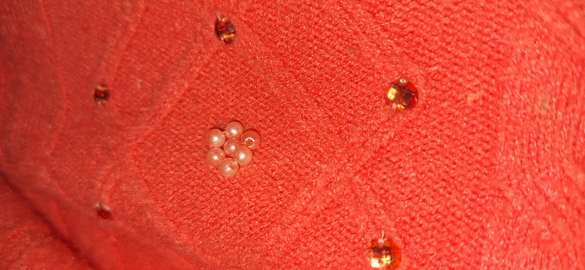 The image is a close-up, overhead shot of a textured, coral-red fabric. Scattered across the fabric are several small, light-pink, iridescent beads arranged in a flower-like pattern in the center, along with three reddish-orange, faceted embellishments. The fabric has a fuzzy, knit texture, and the lighting suggests it might be artificial. There is no discernible location in Bila Tserkva, Ukraine, or any people, actions, or interactions depicted.