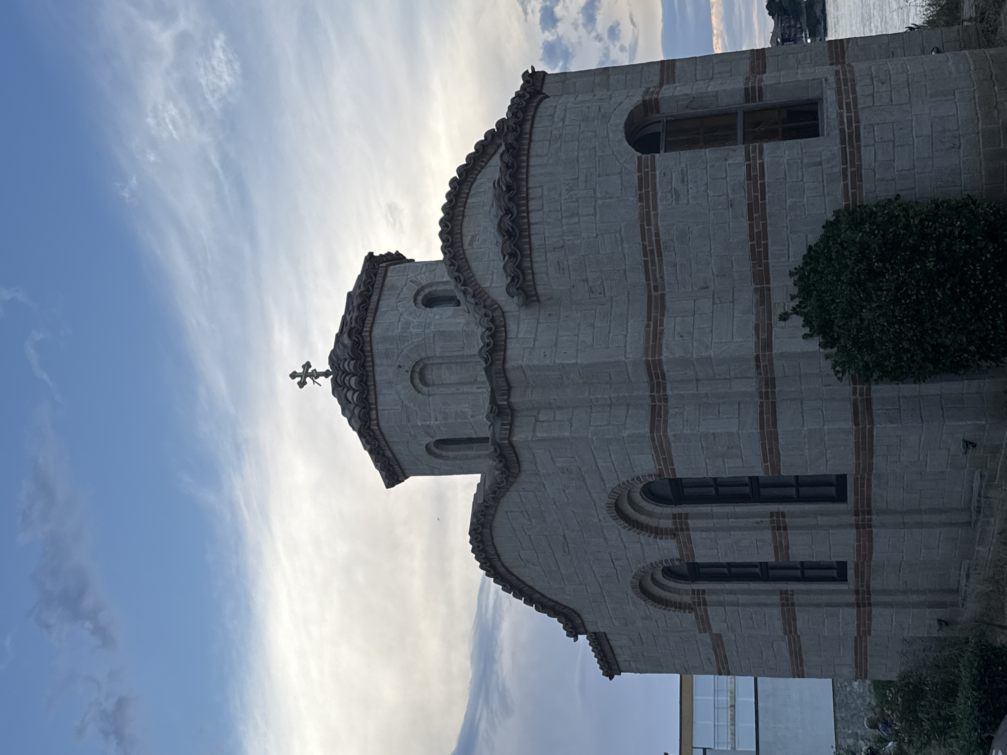 A low-angle, full shot captures the exterior of a stone church with a tiled roof and a cross on top. The church is constructed from light gray stone blocks with horizontal bands of reddish-brown brick. It features arched windows and a domed central section. Green bushes are visible in the foreground, and a building with white walls and a blue-tinted window is partially visible to the left. The background shows a body of water and a hazy sky with clouds. This is the Church of Saint John the Baptist, located in Sozopol, Bulgaria.