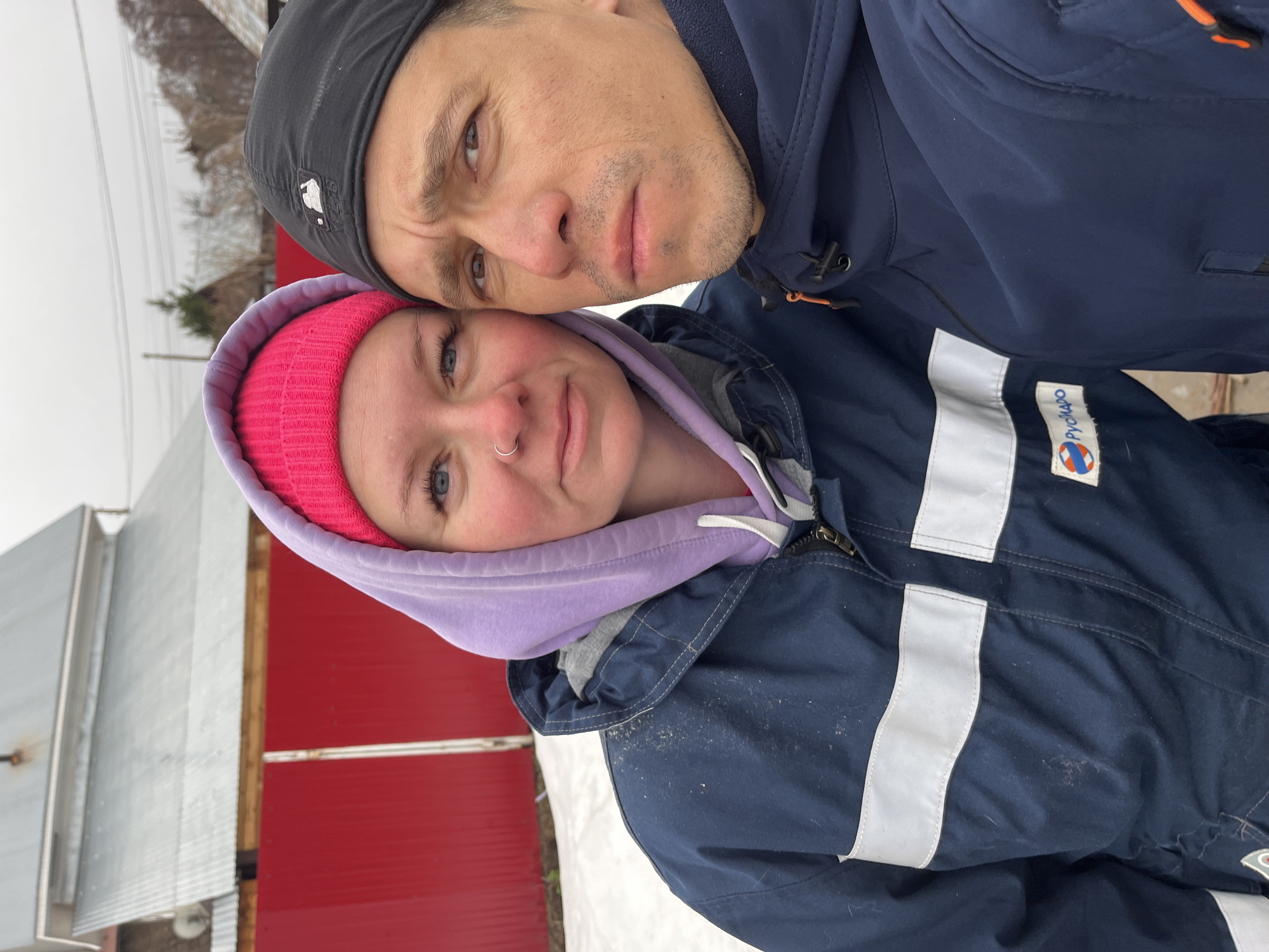 A selfie picture depicts a man and a woman standing close together outside. The man, wearing a black baseball cap and a dark blue jacket, has his face near the top of the frame. The woman, wearing a pink beanie and a purple hooded sweatshirt under a dark blue jacket with white stripes, is positioned below him. In the background, there is a red metal building and a snowy environment. The jackets they are wearing have "РусГидро" logos on them, indicating that this photo was taken within the city of Chaikovsky, Russia.