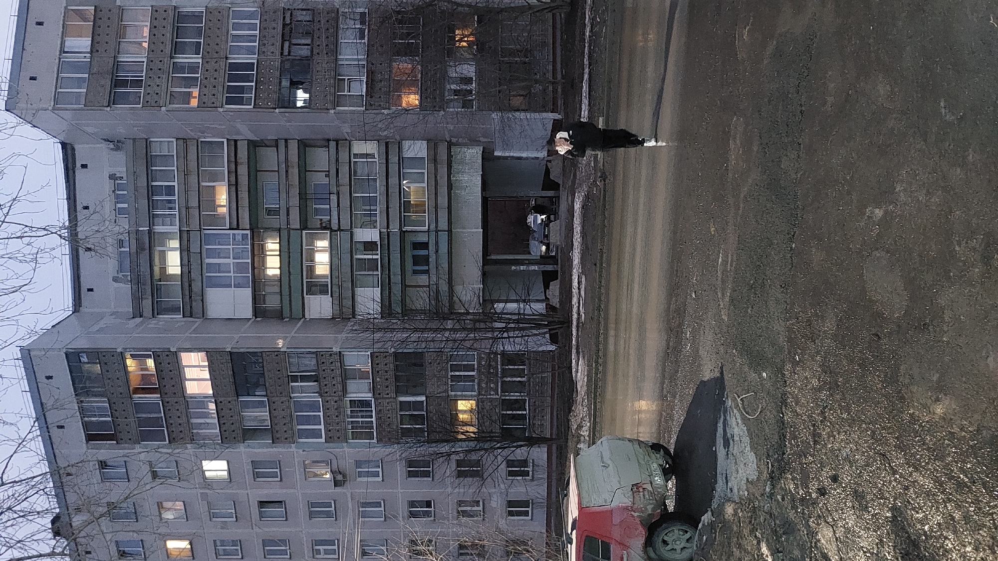 The scene is an urban landscape in Novosibirsk, Russia. A tall, grey apartment building dominates the left side of the frame, with numerous windows, some illuminated. Bare tree branches are visible in front of the building. A person wearing a dark jacket and light-colored mask stands on a wet street, possibly a pedestrian. A partially visible red vehicle is parked on the right, with its side damaged and covered with gray tarpaulin. The street surface appears wet and dark, suggesting recent rain or snow melt. The sky is overcast and grey, indicating a gloomy weather condition.