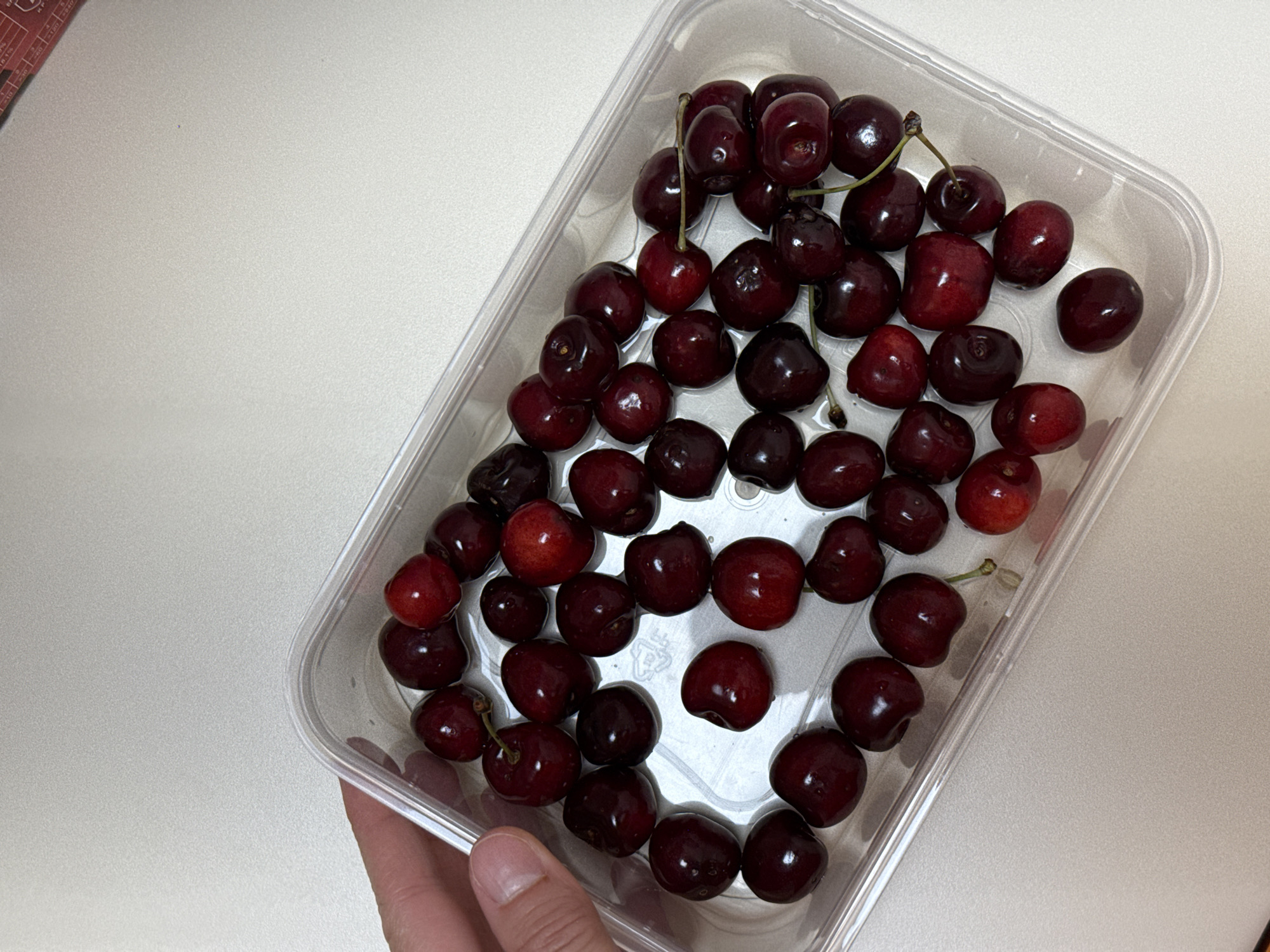 The image shows a clear plastic container filled with dark red cherries. The cherries are plump and glossy, with some still attached to their stems. The container is placed on a light-colored, textured surface, possibly a table or countertop. A hand is visible in the lower left corner, holding the container. The scene appears to be indoors, with soft, diffused lighting. There is no text visible in the image. The overall impression is a simple, everyday scene of fresh fruit.