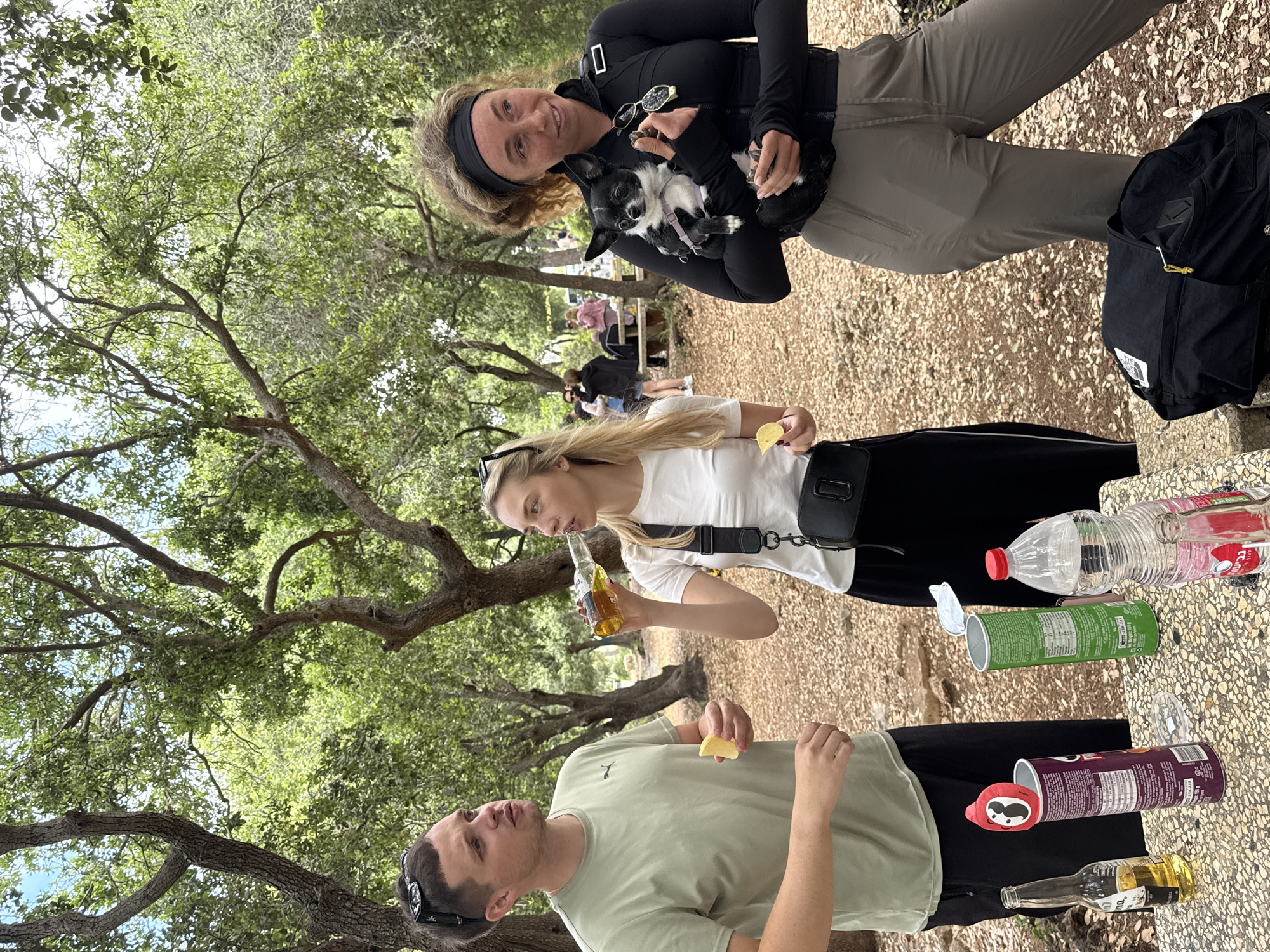 Nesher, Israel. A group of three individuals are gathered outdoors at a picnic table. One individual is holding a small dog. Two individuals are holding slices of lemon. One individual is drinking from a bottle. An opened container of Pringles potato chips is on the table.