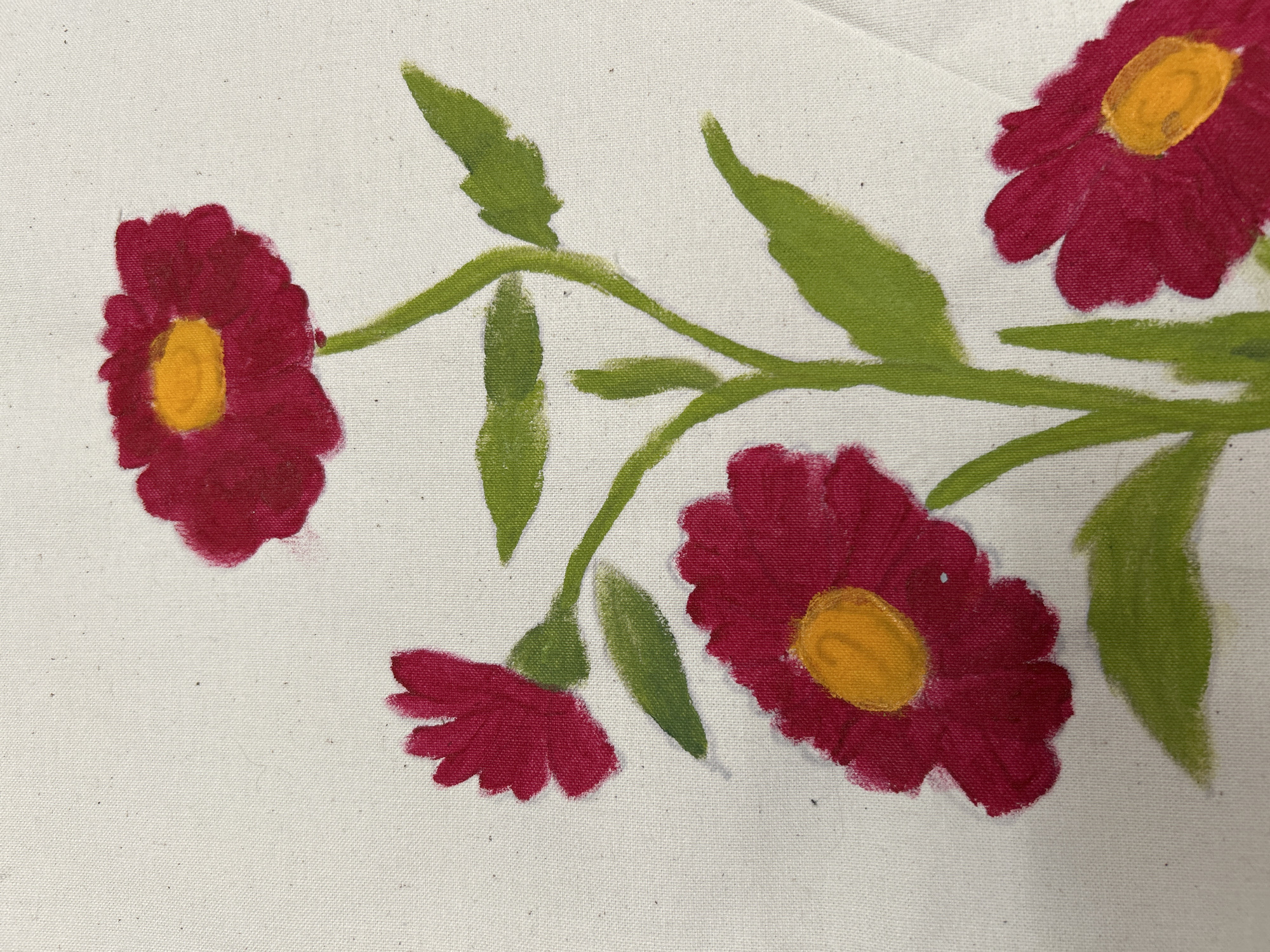 A close-up shot presents a painted floral design on a white canvas-like material. The design features four red flowers, each with a yellow center. Green stems and leaves connect the flowers, creating a branch-like arrangement. The paint appears thick, with visible brushstrokes. The setting appears to be indoors with plenty of natural lighting.