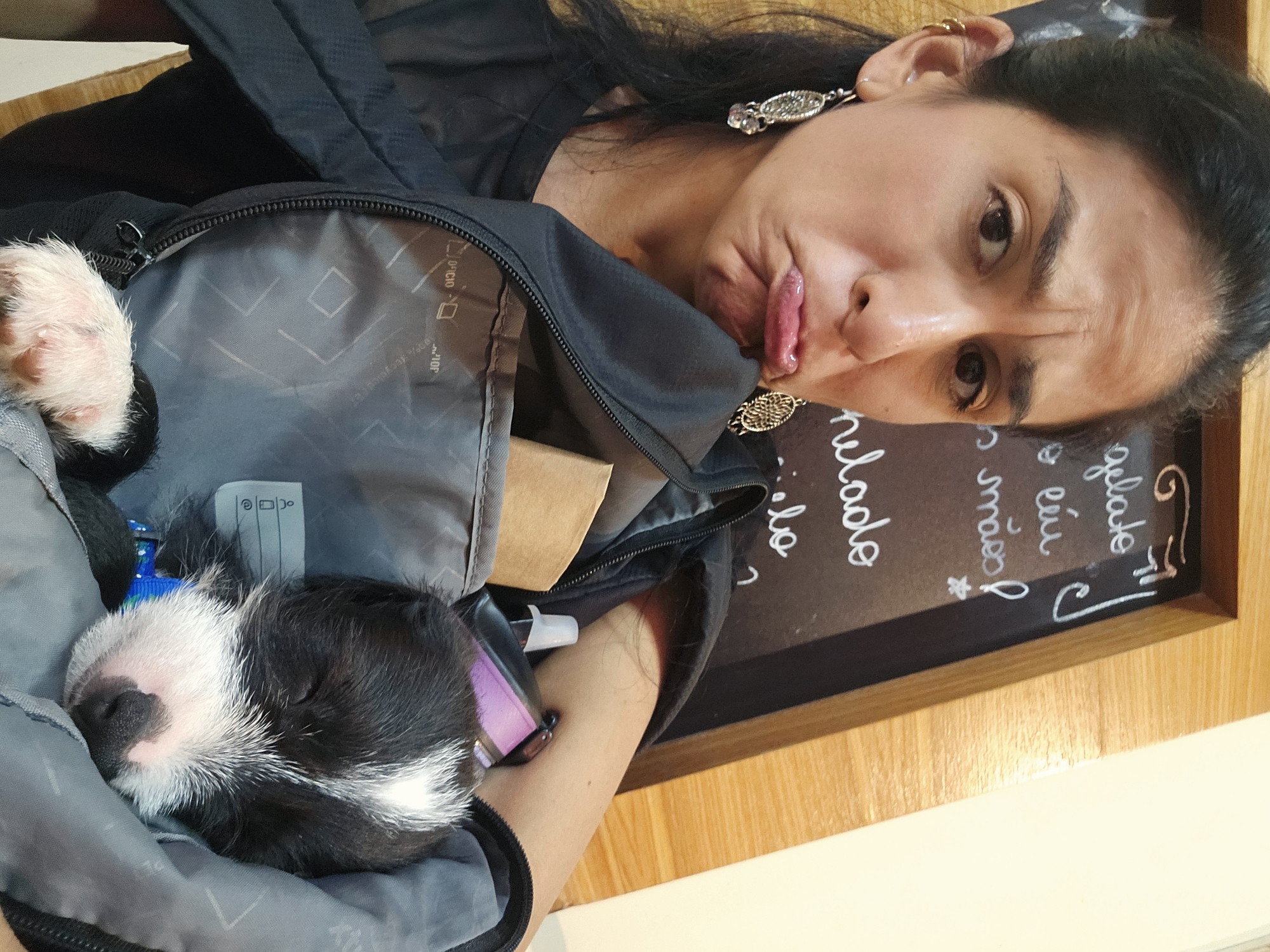 A woman is posing with a puppy sleeping inside of a backpack. The woman has dark hair, earrings, and is making a funny face. The puppy is black and white. A menu written in Portuguese is visible in the background, which translates to "gelato." The location is likely a cafe or restaurant in Armação Dos Búzios, Brazil.