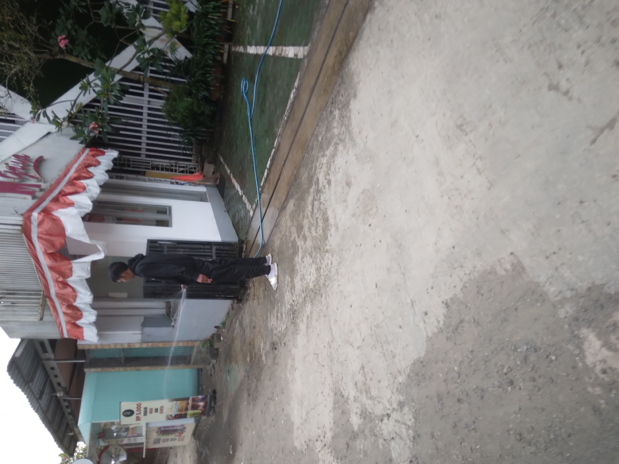 The image shows a person wearing a black hoodie and black pants, standing on a wet concrete surface. The person is holding a blue hose, and water is spraying from it. To the left of the person is a small building with a red and white awning. A sign with the text "RP 5.000" is visible on a kiosk to the left of the building. Behind the building, there is a fence and some greenery. The scene appears to be outdoors, possibly in front of a shop or a residential area in Desa Pamijen, Kecamatan Sokaraja, Indonesia. The ground is damp, suggesting recent rain or watering. The lighting is overcast.