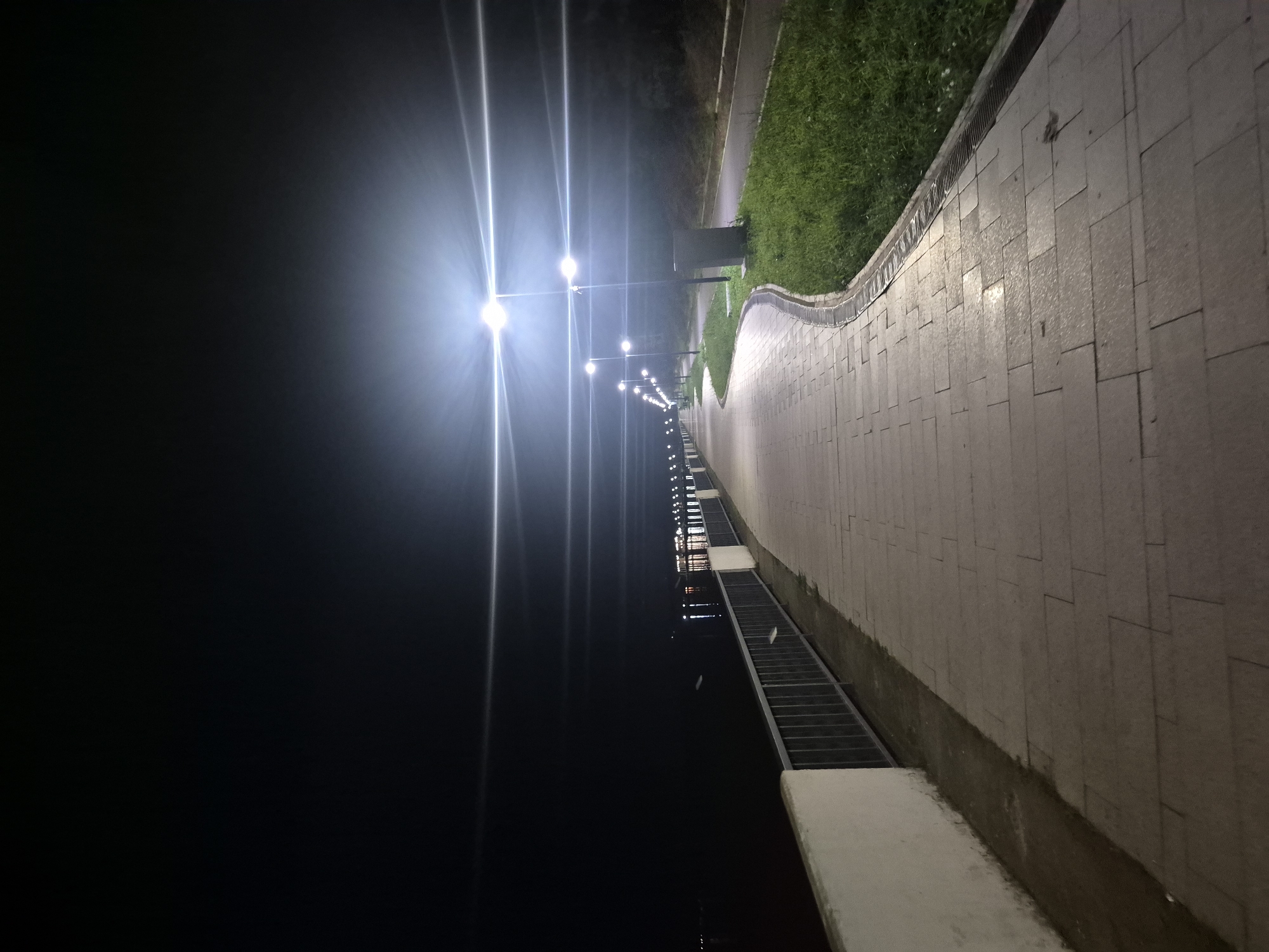 A night-time, vertical, long shot captures an empty, illuminated walkway bordering a dark body of water in Chaikovsky, Russia. The walkway is paved with rectangular stone tiles and is lined with a low, white concrete barrier topped with a metal railing on the water side. Along the opposite side of the walkway, a strip of green grass is illuminated by a row of tall lampposts. The lampposts are spaced evenly apart, casting bright, star-like flares across the dark sky. In the distance, across the water, a line of lights suggests buildings or structures. The overall impression is of a serene and empty urban landscape at night.