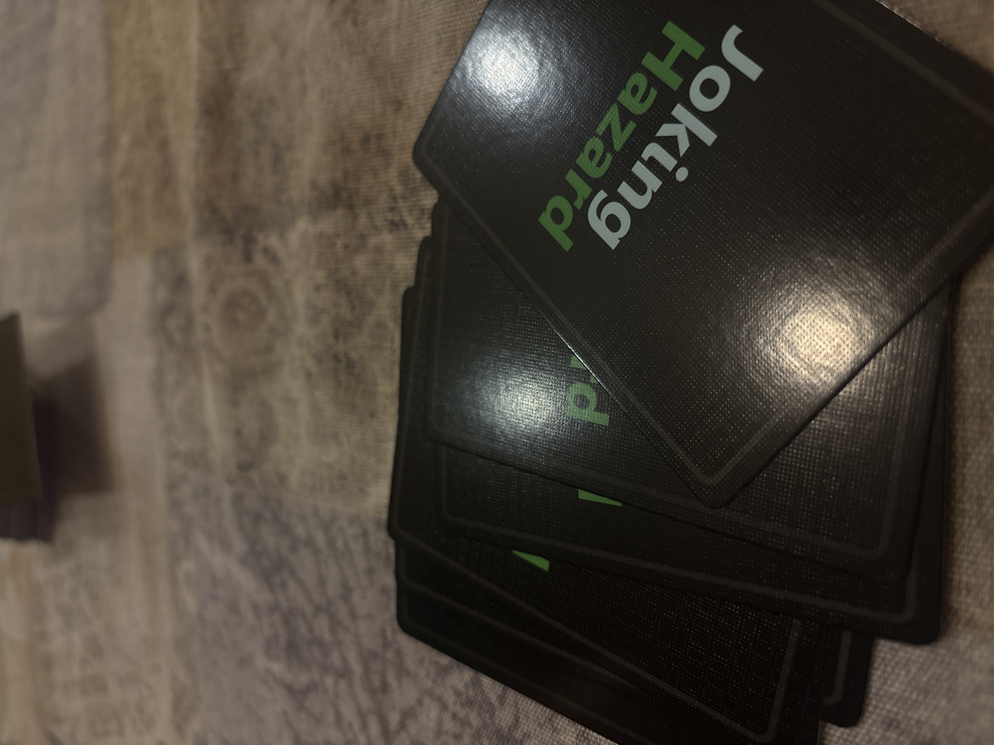 A stack of playing cards from the game "Joking Hazard" is prominently featured in this close-up, angled shot. The cards are black with a noticeable textured finish, evident across their surfaces. The top card, slightly fanned out from the rest, clearly displays the game's title. "Joking" is printed in white capital letters, while "Hazard" is in a contrasting bright green, also in capitals. A strong glare from an unseen light source reflects off the upper left corner of this top card, creating a bright highlight.
Beneath the leading card, several other black cards are partially visible, layered slightly askew, revealing hints of the same green color, likely from the "Hazard" text on their backs. The background consists of a soft-focus, textured surface, possibly a patterned rug or tablecloth, in varying muted tones of brown, beige, and grey. No people or specific activity are depicted, suggesting the cards are either at rest before or after a game. The setting is indoors, with no distinct location cues other than the provided context of Plovdiv, Bulgaria. The image captures the details of the card backs and the immediate playing surface.