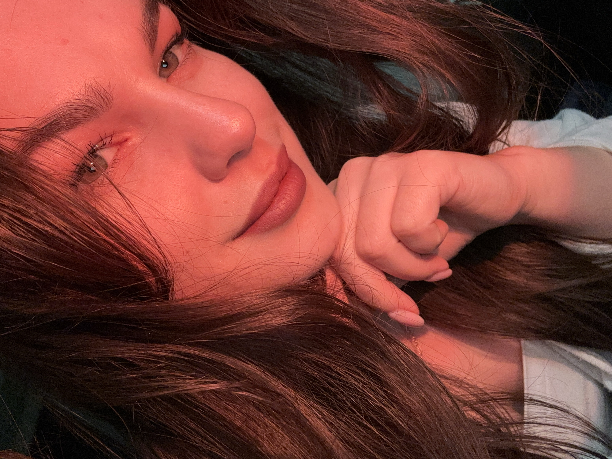 The image shows a close-up of a woman's face, illuminated by a reddish light. She has brown hair, fair skin, and hazel eyes. Her hand is near her face, resting under her chin. She has lip color on her lips. The location in the city of Chaikovsky, Russia, is not verifiable from this image.