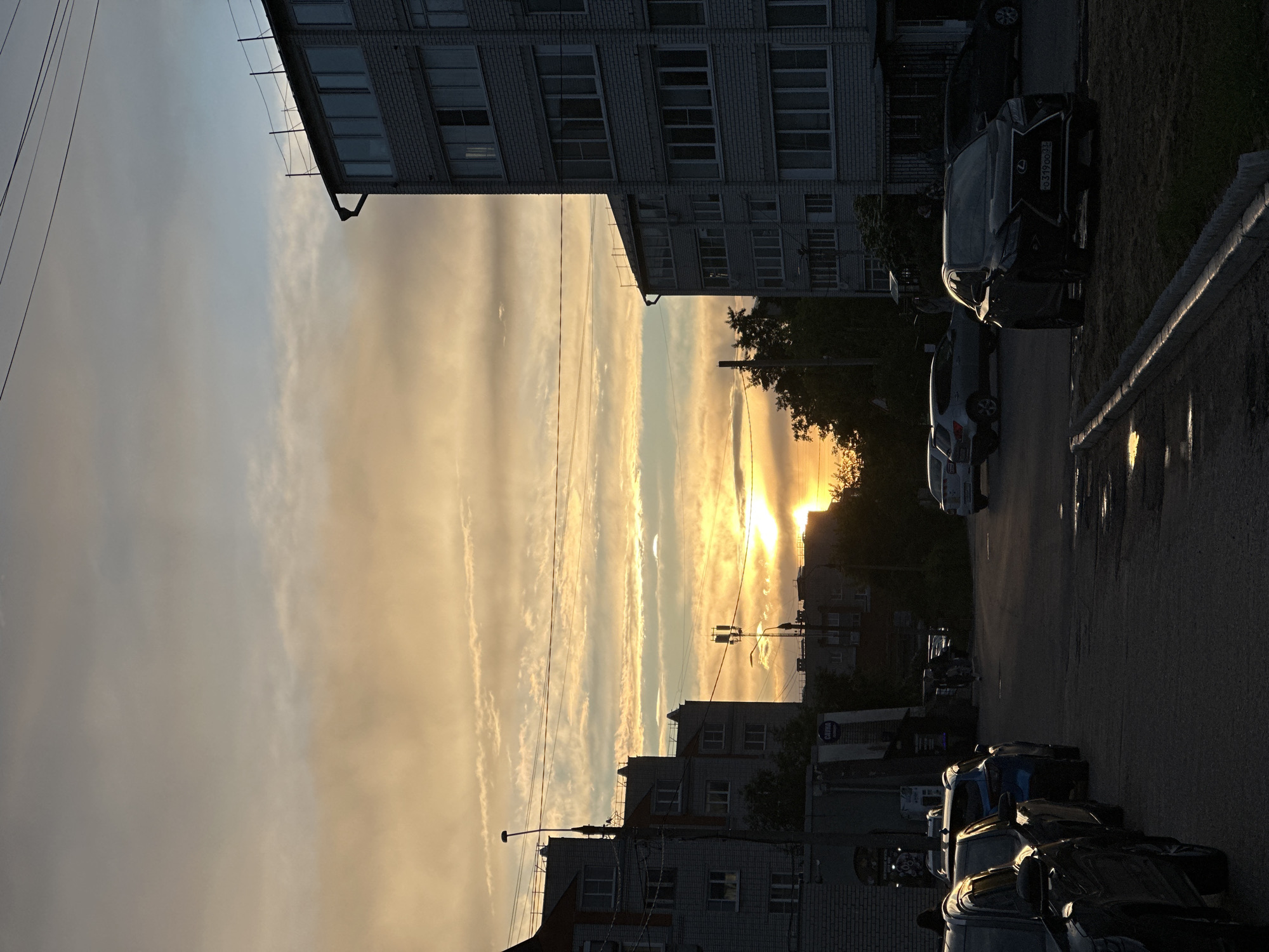 The image depicts an urban sunset scene in Vladimir, Russia. A multi-story brick building is visible on the left, with rows of rectangular windows. Power lines run diagonally across the sky. In the distance, the sun sets between buildings, creating a bright orange glow. Cars are parked along the right side of the street, partially obscured by shadows. The asphalt street is dark, with reflections visible. The sky is filled with streaky clouds, colored with golden and grey hues. A few trees and buildings are silhouetted against the setting sun. The image appears to be taken from street level, with a slight upward angle.