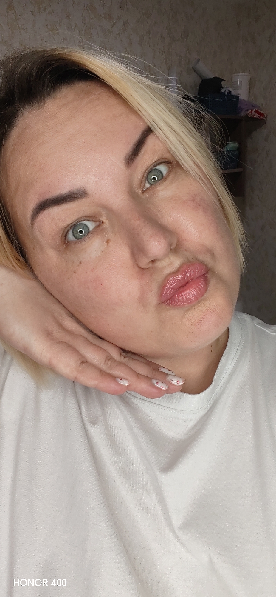 A close-up shot shows a woman with blonde hair, fair skin, and light green eyes, pursing her lips in a kiss-like gesture. Her left hand is raised to her cheek, displaying short nails painted white with small red floral designs. She is wearing a white t-shirt. In the blurred background, a shelf with various objects is visible. The bottom of the frame shows the text "HONOR 400." This image does not provide sufficient information to establish a location in the city of Noviy, Russia.