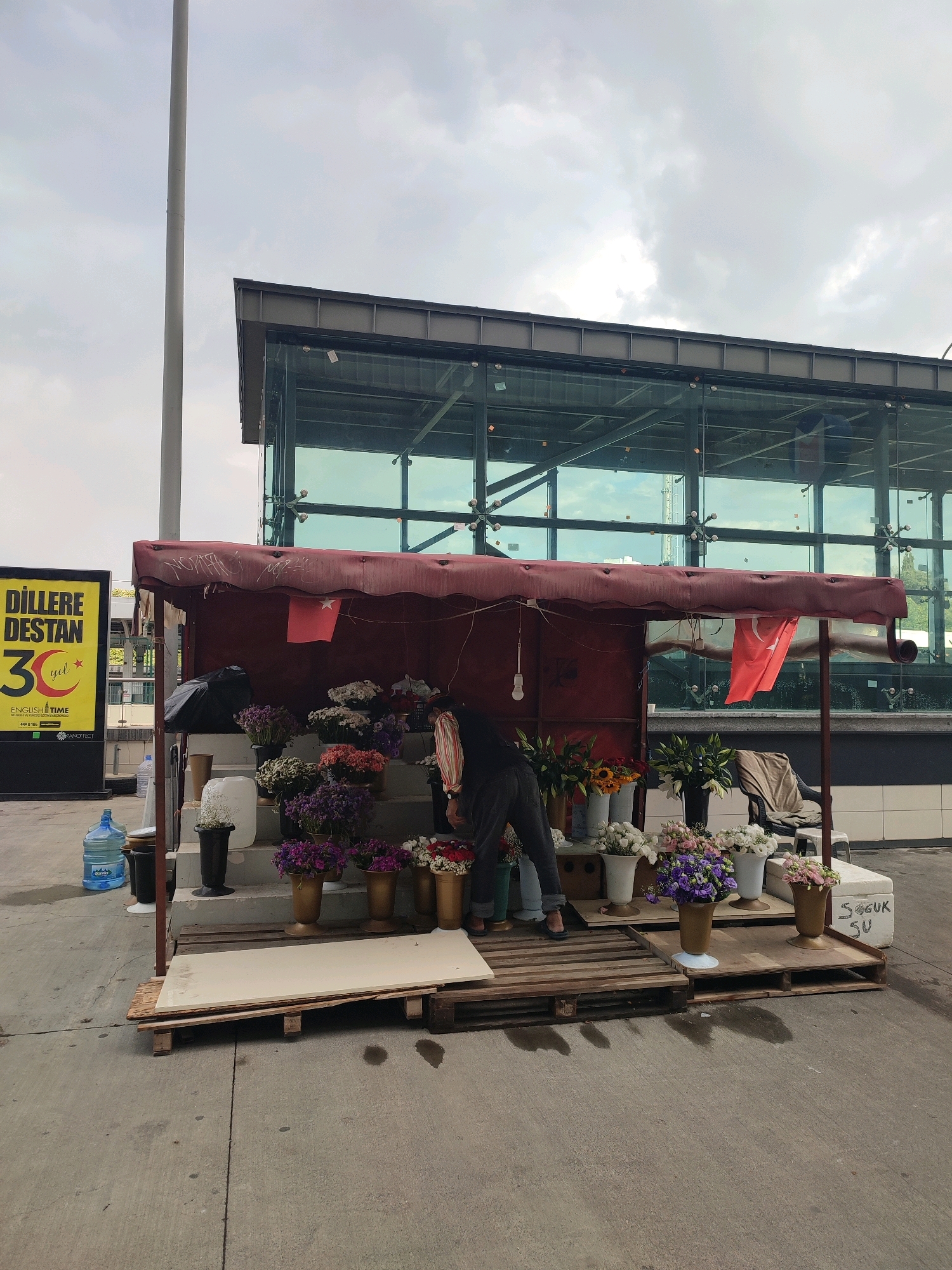 The image depicts a street scene in Kadıköy, Turkey, featuring a flower stall set up on wooden pallets in front of a modern building with a glass facade. A person, dressed in dark clothing and jeans, is attending to the flowers. The stall has a burgundy awning and displays various potted flowers in vases, including purple, pink, and white varieties, as well as some sunflowers. A Turkish flag is visible hanging from the awning. To the left of the stall is a large yellow advertisement sign with Turkish text. In the background, the building appears to be a commercial or public structure with large windows reflecting the overcast sky. Several large water jugs are also present near the stall. The ground is paved concrete.
