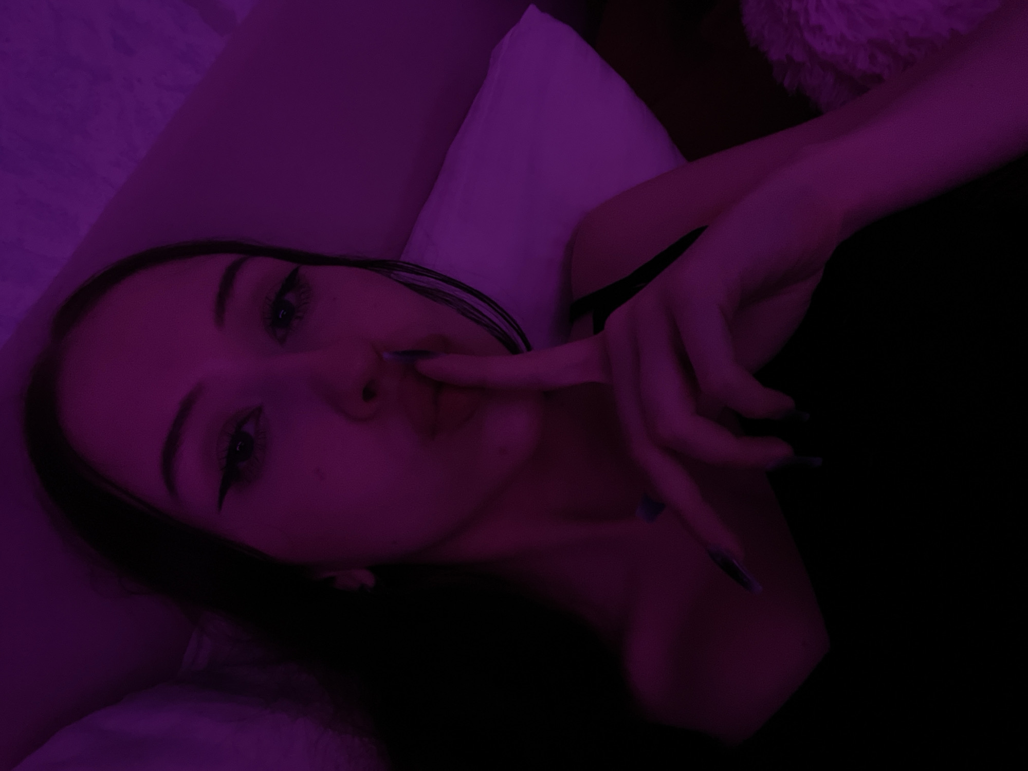A young woman with dark hair and winged eyeliner is lying down, illuminated by purple light. She has her index finger to her lips in a "shushing" gesture, her mouth slightly open in a kiss pose. Her nails are long and dark. She is wearing a black top. The background appears to be a bed with a white pillow and a textured surface that may be a blanket or rug. The image is a close-up, and the woman's face and upper body are the primary focus. There is no identifiable information about the location, including Votkinsk, Russia.