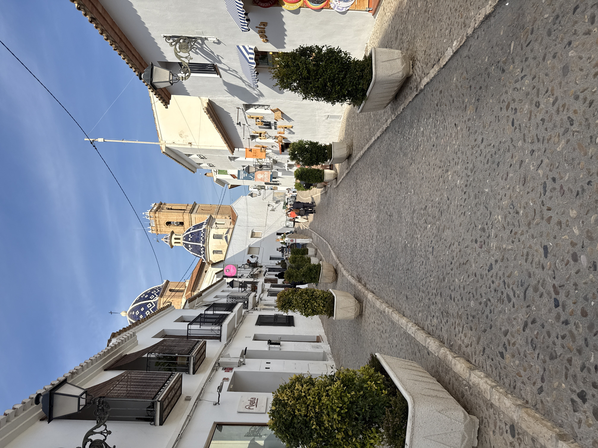 A low-angle, eye-level, full outdoor shot depicts a narrow street in Altea, Spain, angled to the right. The street, made of cobblestones, is flanked by white buildings on both sides. The buildings are adorned with potted plants, balconies, and awnings. In the distance, a church with a blue-tiled dome stands out. A few pedestrians can be seen walking along the street. The sky is clear and blue. Overhead, a power line runs along the length of the street.