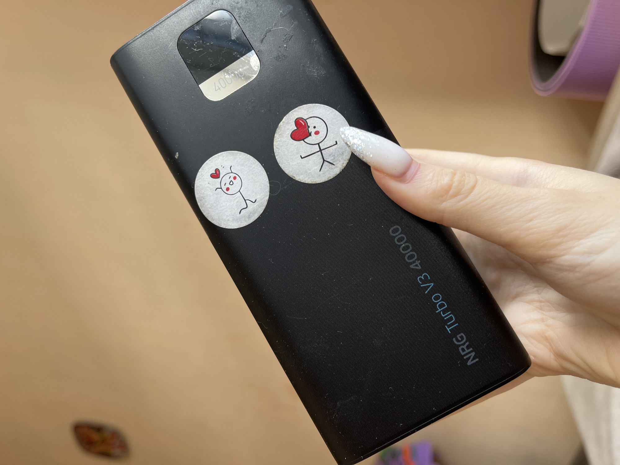 A person's hand, with a long, white, glittery fingernail, is holding a black power bank. The power bank has two circular stickers with cartoon drawings of stick figures with red hearts above their heads. Text on the power bank reads "NRG Turbo V3 40000". A small, blurred object with orange and yellow coloring is visible on the wall in the upper left of the frame. The background is a plain, light brown wall. The image does not provide any information about the city of Ivanovo, Russia.