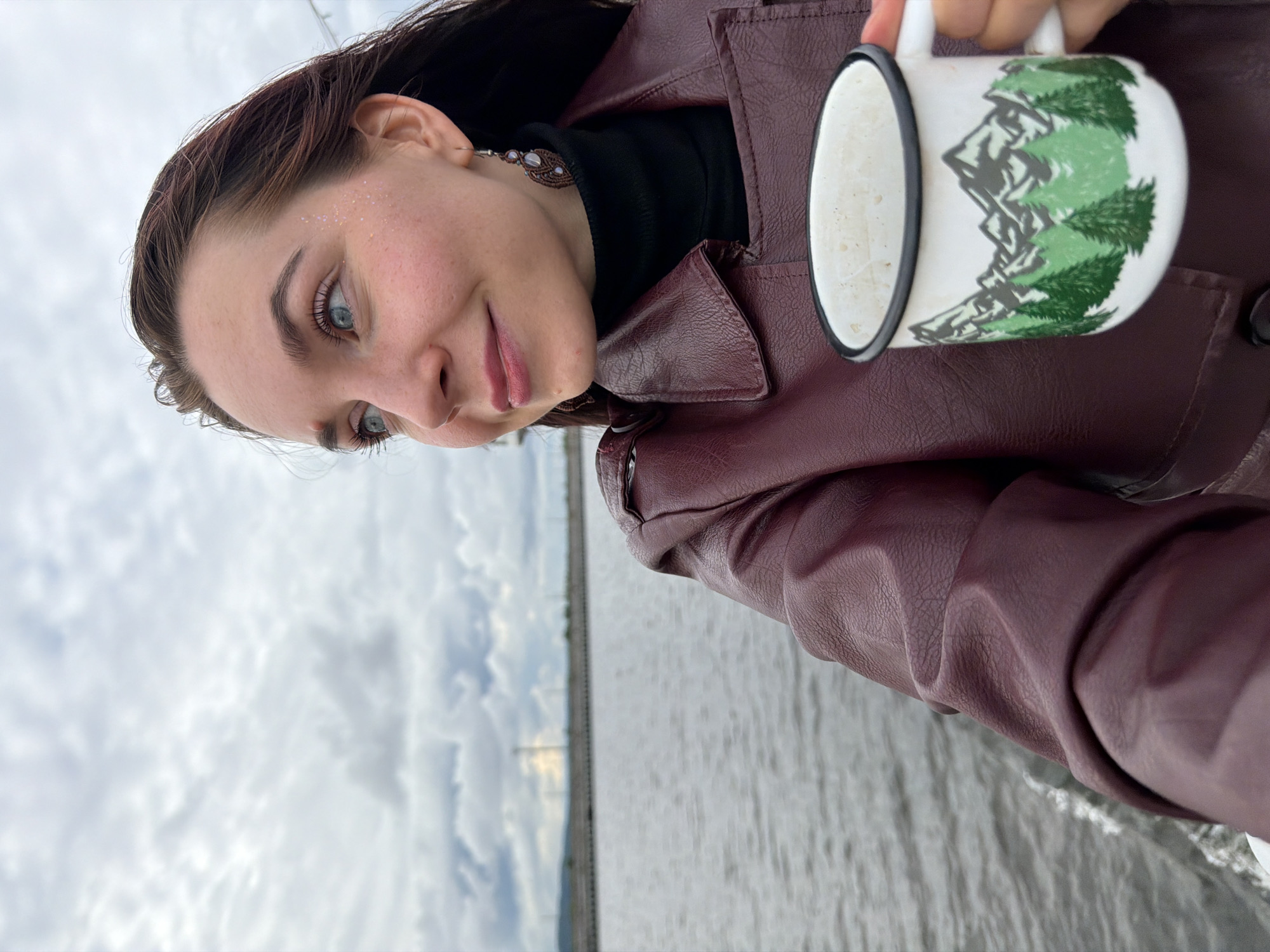 A young woman with brown hair and blue eyes is holding a white enamel mug with a painted landscape of mountains and green trees. She is wearing a burgundy leather jacket and a black turtleneck. Her face is turned to the side, and she is smiling slightly. The background shows a body of water with a cloudy sky overhead. The photo appears to have been taken outdoors, possibly on a boat, as indicated by the movement of the water. The location is identified as Chaikovsky, Russia.