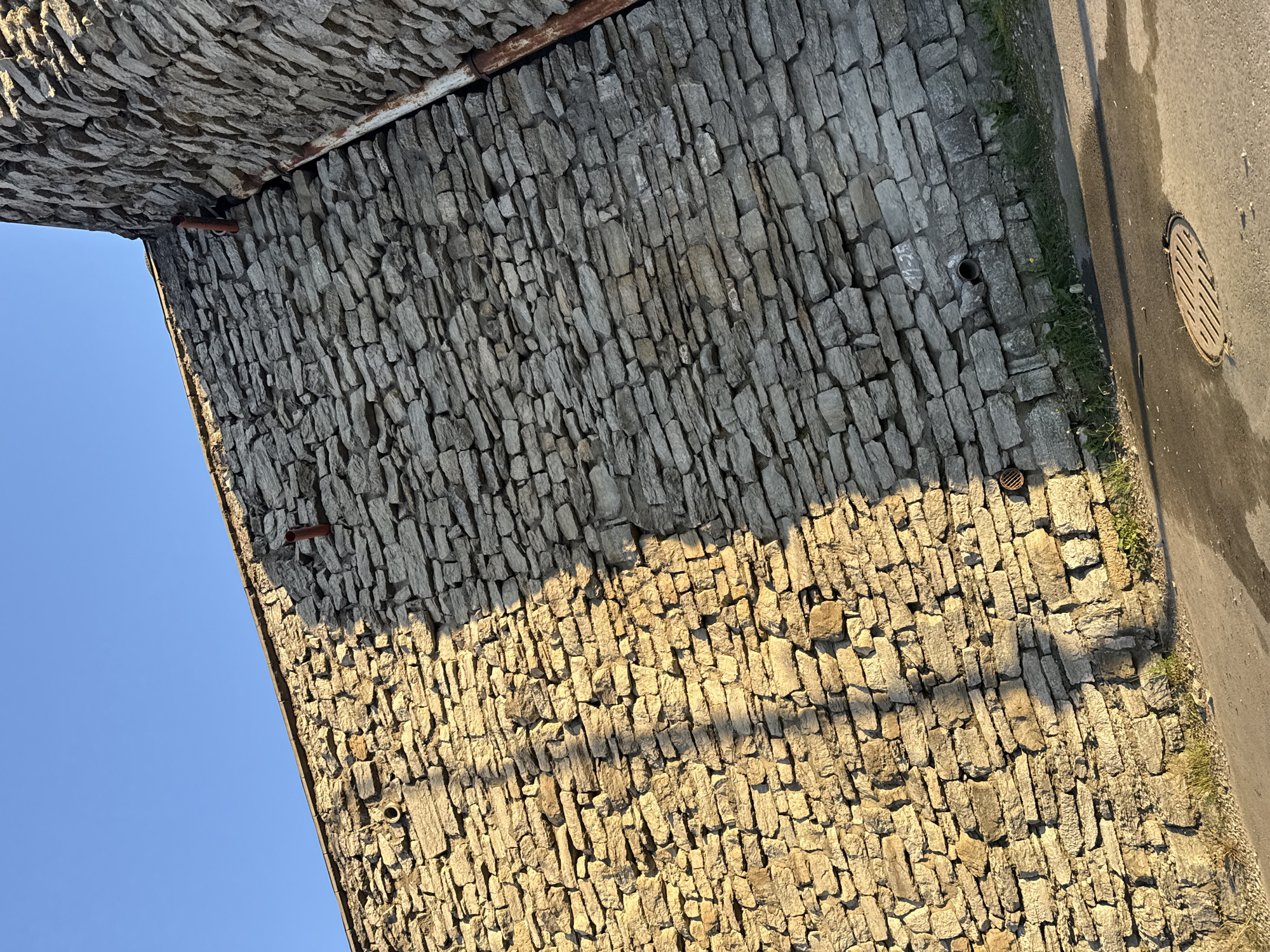 The image shows a close-up view of a stone wall in Tallinn, Estonia. The wall is constructed from irregular, grey stones and is partially illuminated by the warm light of the setting sun, casting long shadows. A section of the wall is darker due to a shadow, suggesting the presence of an object or structure casting it. Several small, round openings are visible in the wall, possibly for ventilation or drainage. A rusty metal pipe runs vertically along the corner of the wall on the right side of the image. In the foreground, a wet asphalt surface is visible, with a manhole cover partially submerged in the water. The sky above is a clear, deep blue. The overall impression is one of old, robust architecture bathed in the golden hour light.