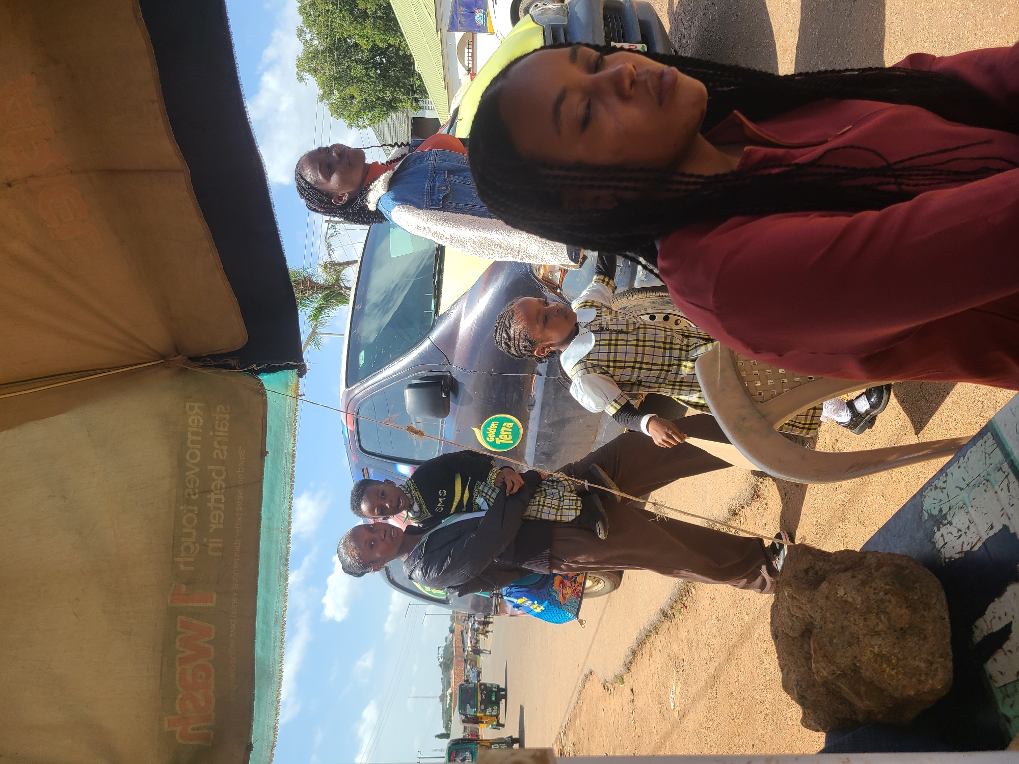 This media file shows an outdoor scene in Jos, Nigeria, under a clear, sunny sky, suggesting midday. The image features several people, primarily young women and children, gathered around a vehicle. One young woman is prominently in the foreground, with her eyes closed, possibly resting or sleeping, her long braided hair cascading down her shoulder. She is wearing a maroon jacket. Behind her, another young woman in a denim jacket with a fluffy white collar looks towards the camera.
A child is being held by another person, and both are looking at the camera. The child is wearing a checkered shirt. Another person, also dressed in a checkered shirt, is seated on a white plastic chair, their legs crossed, wearing black shoes and white socks. A large, rough rock is positioned on the ground in the lower right corner of the image, partially in shadow.
In the background, a car with a "Golden Terra" sticker is visible, along with other vehicles, including what appear to be auto-rickshaws, hinting at a busy roadside environment. An umbrella or canopy extends from the left side of the frame, casting a shadow and providing some shade. The ground appears to be a dusty, unpaved road. The overall atmosphere seems relaxed, with the subjects appearing comfortable and unconcerned. There is visible text on the umbrella: "How I Wash" and other illegible text. The "Golden Terra" sticker on the car is also readable.