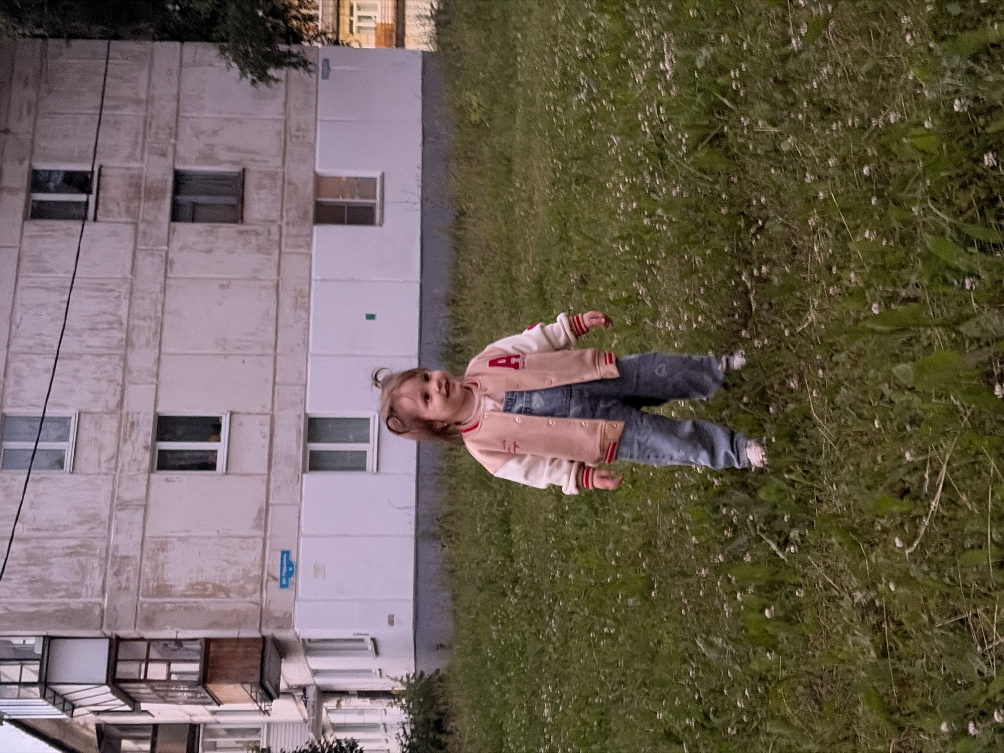 A young child with blonde hair tied in two pigtails stands in a grassy field in front of a multi-story apartment building in Chaikovsky, Russia. The child is wearing a beige and pink varsity jacket with a red stripe on the cuffs and collar, and blue denim overalls. The child is smiling and looking to the right. The apartment building appears to be made of concrete panels and has several windows. A blue street sign is visible on the side of the building. The grass is green and dotted with small white flowers. Trees are visible in the background.