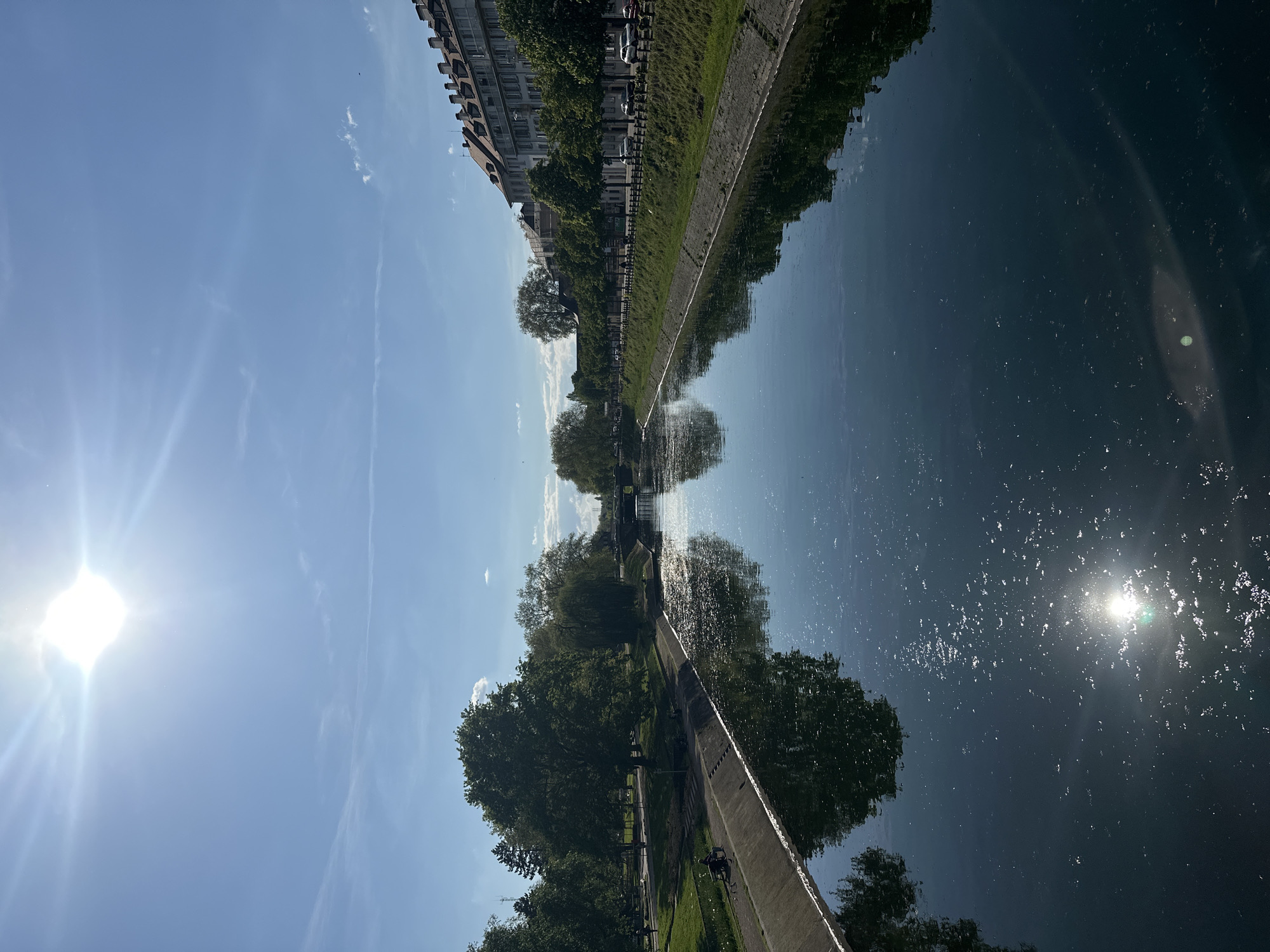 This is an outdoor image depicting a canal reflecting a sunny sky and surrounding urban landscape, likely taken in Strasbourg, France. The orientation is rotated 90 degrees clockwise. The canal runs horizontally, with buildings and trees visible along its banks, forming reflections in the water. The sky is blue with a visible sun glare in the top right and left, creating a sharp contrast. People appear to be walking or cycling along a path beside the canal. The overall environment suggests an urban green space within the city.