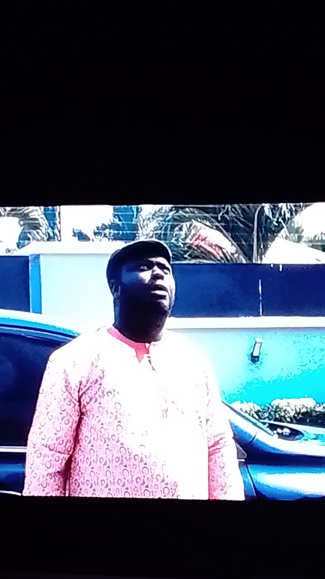 A medium shot captures a Black man wearing a flat cap and a pink patterned shirt, looking upwards. He is positioned in front of a blue and light-colored wall, with a car visible to his left. The background features foliage and a bright blue sky. The man has dark skin and is looking upwards with his mouth slightly open. His shirt has a repeating circular pattern in a darker pink.