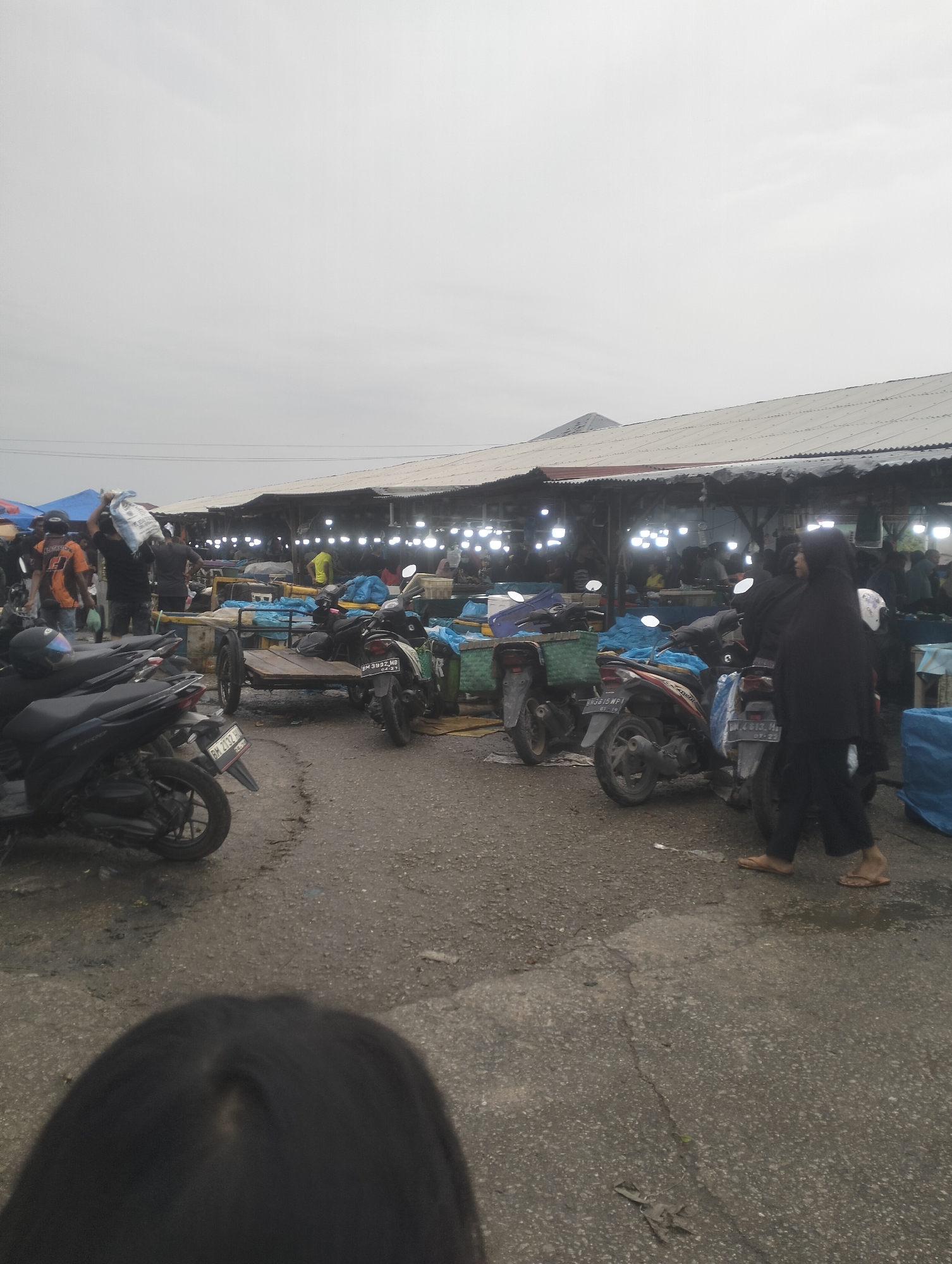 The image depicts an outdoor market scene in Dumai City, Indonesia, likely a fish market based on the presence of blue tarps and the overall atmosphere. The market is covered by a long, corrugated metal roof supported by wooden posts, with numerous exposed light bulbs illuminating the stalls. In the foreground, several motorcycles are parked on a paved surface that appears to be asphalt with visible cracks and puddles, suggesting recent rain or damp conditions.
Several people are present in the scene. On the left, a person in an orange and black shirt is carrying a white bag over their shoulder. To the right of the motorcycles, a woman in a black hijab and dark clothing is walking, wearing flip-flops. Other individuals are visible in the background, milling around the stalls under the covered market area. Some are seated or standing behind counters, presumably vendors. A wooden cart with two wheels is also positioned in the market. The sky is overcast, contributing to a diffused light. The scene conveys a bustling, everyday market environment.