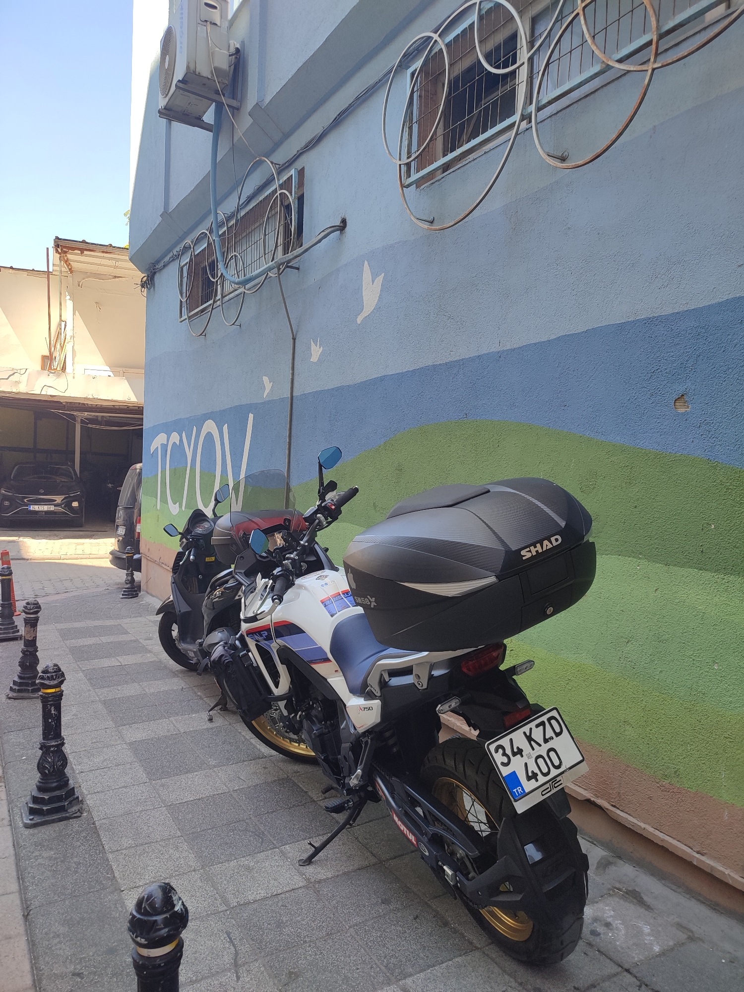 The image depicts an outdoor scene on a city street, likely in Kadıköy, Turkey, given the Turkish license plate. The focal point is a white and blue adventure-style motorcycle parked on a paved walkway. The motorcycle has a black Shad top case and a visible license plate that reads "34 KZD 400 TR". To the left of the motorcycle, a dark-colored scooter is also parked. In the background, a building with a light blue and green mural is visible. The mural features abstract shapes and the word "TCYOV" in large white lettering. An air conditioning unit is mounted high on the building wall. A partial view of a parking garage entrance with a black car inside is visible on the far left. The walkway is lined with decorative black bollards. The lighting suggests it is daytime.