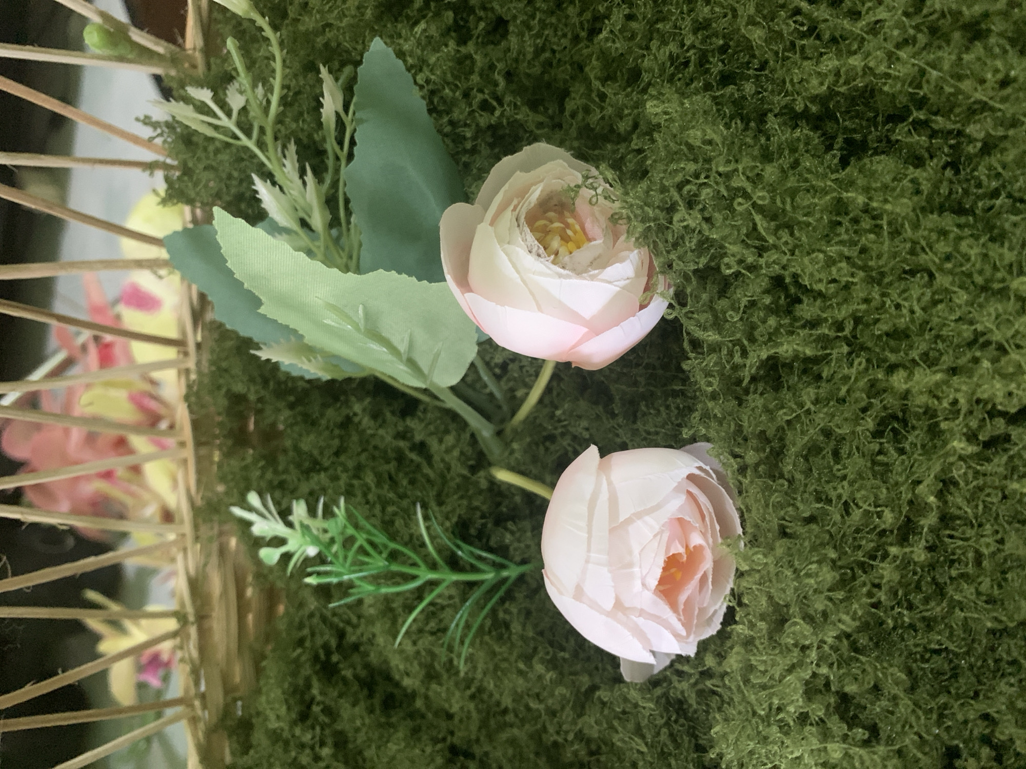 A close-up shot captures two light pink peonies nestled in a bed of fluffy, bright green faux moss. To the left, a delicate sprig of artificial greenery with thin, pointed leaves stands upright. Behind the flowers, a portion of a woven basket is visible, its light brown wooden spokes fanning upwards. In the blurred background, hints of more colorful artificial flowers, appearing to be pink and yellow, are present within the basket. The scene is composed from a slightly overhead angle, with the focus sharp on the peonies and moss. The image does not contain any elements that would suggest a specific location in Kuala Pembuang, Indonesia.