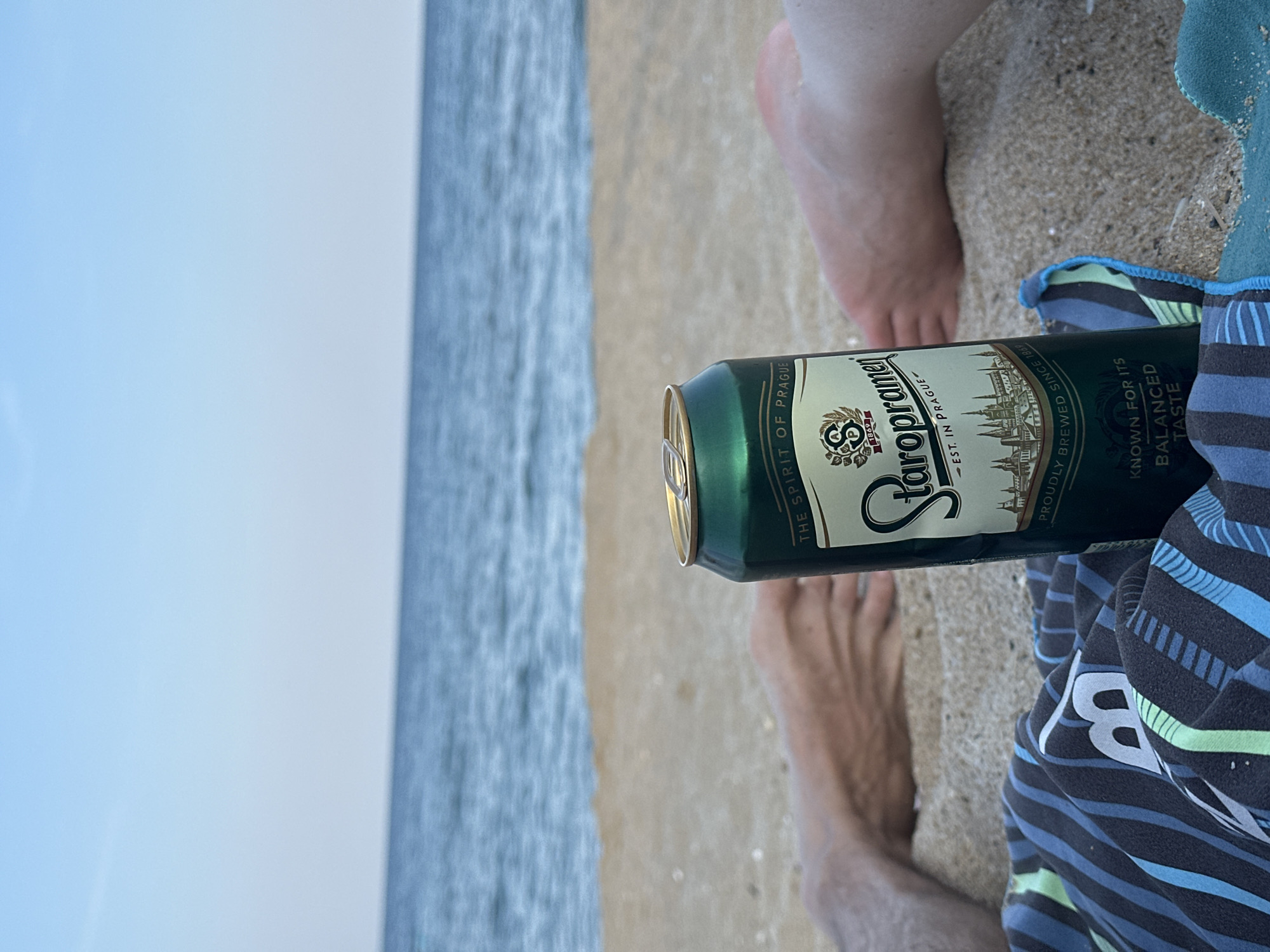 The image captures a close-up, low-angle view of a green can of Staropramen beer on a sandy beach. The can is positioned in the foreground, slightly to the right of the center. Behind the can, two people's bare legs are visible, partially buried in the sand, suggesting they are lounging on the beach. A striped towel is laid out in the lower left portion of the frame. In the background, the calm blue ocean meets a clear sky, creating a serene beach scene. The overall impression is one of relaxation and leisure.