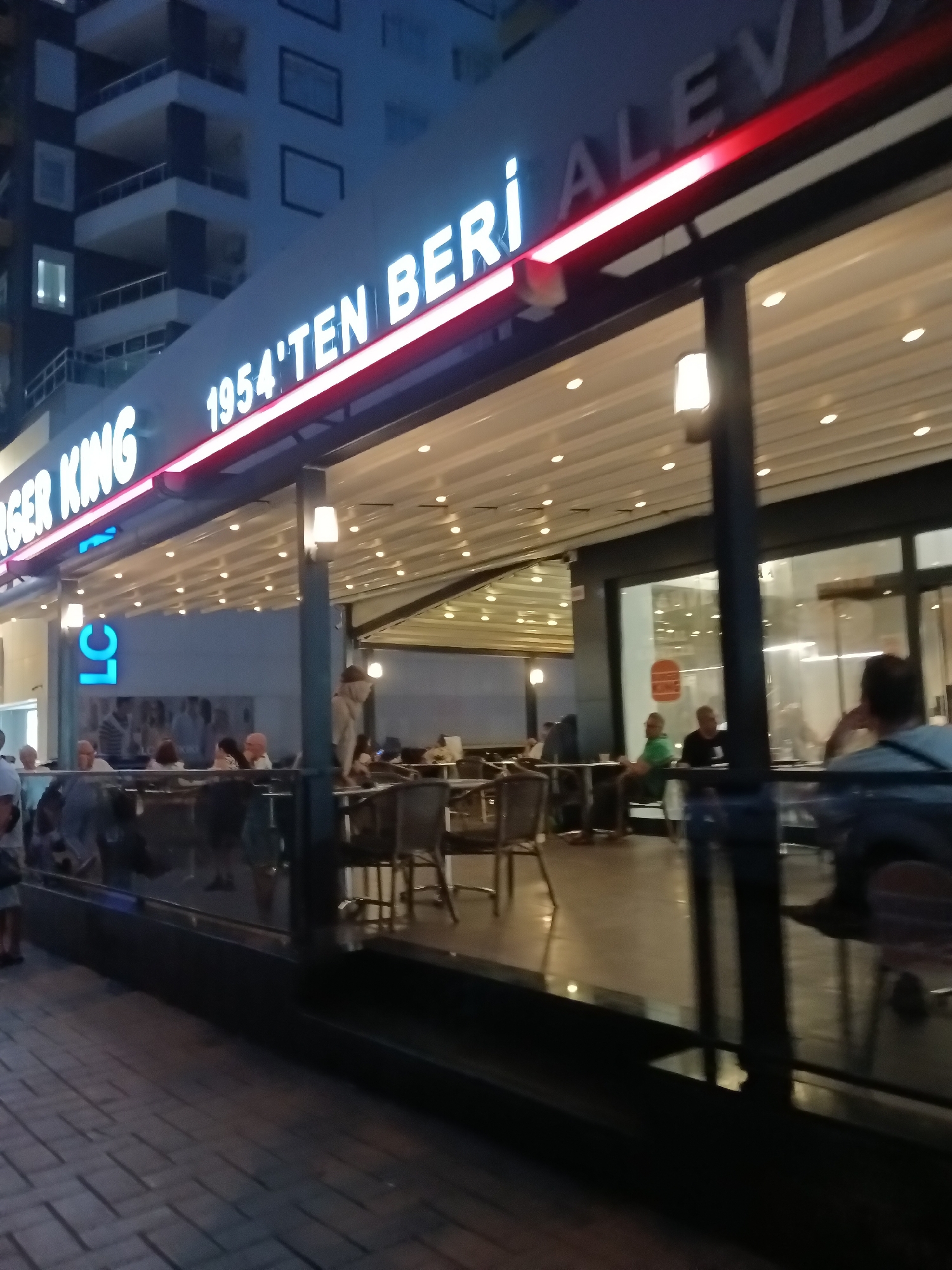 This image shows the exterior of a Burger King restaurant in Alanya, Turkey. The restaurant has a prominent neon sign that reads "BURGER KING 1954'TEN BERİ ALEV". The restaurant is lit with overhead lights, and there are tables and chairs both inside and outside. Several people are visible sitting at tables, some eating and some talking. A building with multiple floors is visible behind the restaurant, and there is a sidewalk in front of the restaurant with a brick-like pattern. The overall scene suggests a busy evening at a popular fast-food establishment.