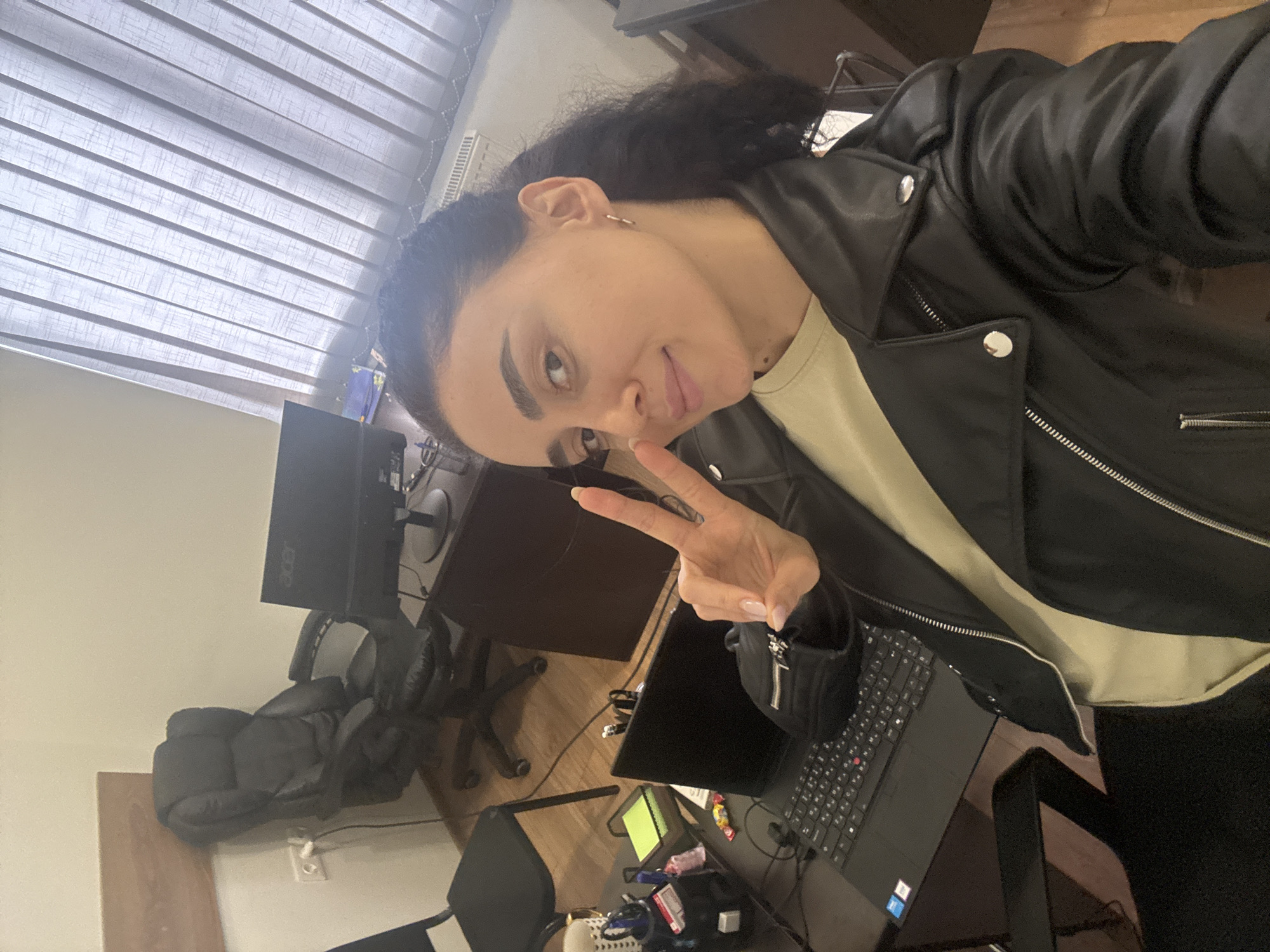 The image shows a person in Tbilisi, Georgia, taking a selfie in an office setting. The person is wearing a black leather jacket and making a peace sign with their right hand. The background features a desk with a computer monitor, keyboard, mouse, and other office supplies. A black office chair is also visible. Light streams in through a window with blinds.